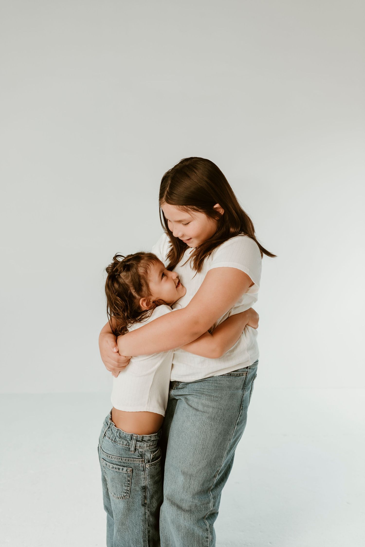 Two sisters sharing a warm hug in a white studio, older girl embracing younger child tenderly.