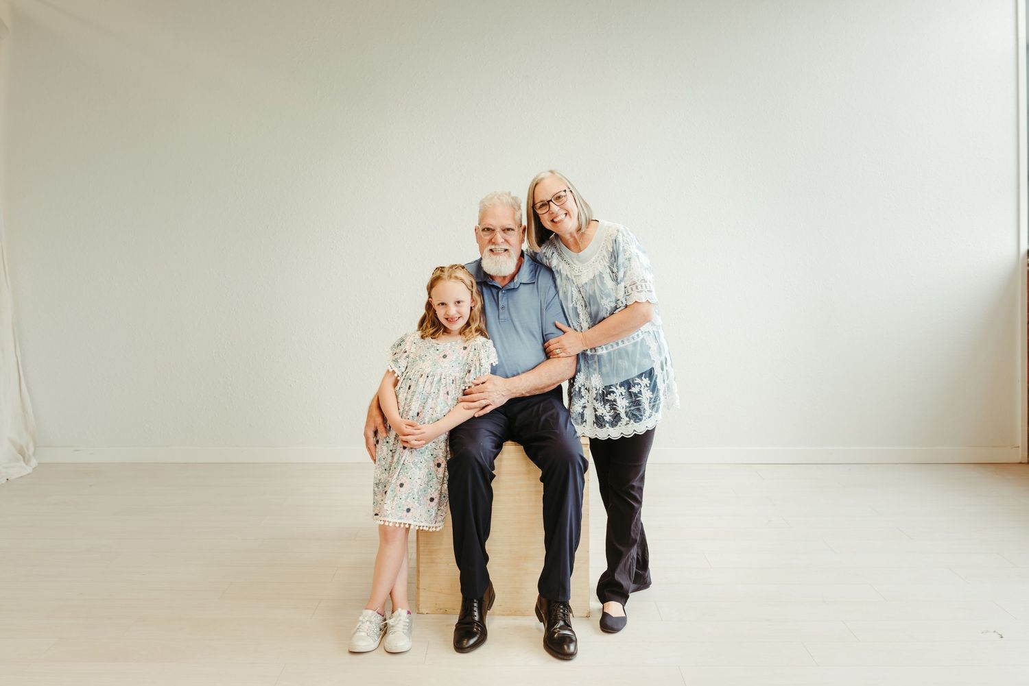 Grandparents posing with young granddaughter in floral dress in bright white studio setting.