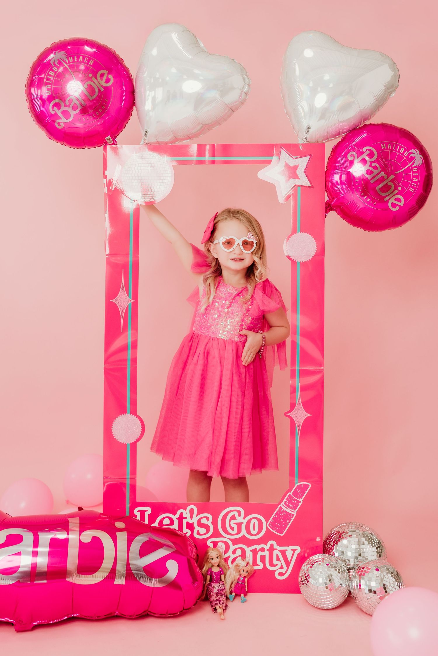 Young girl in hot pink sequin dress posing inside Barbie themed photo frame with balloons on pink backdrop.