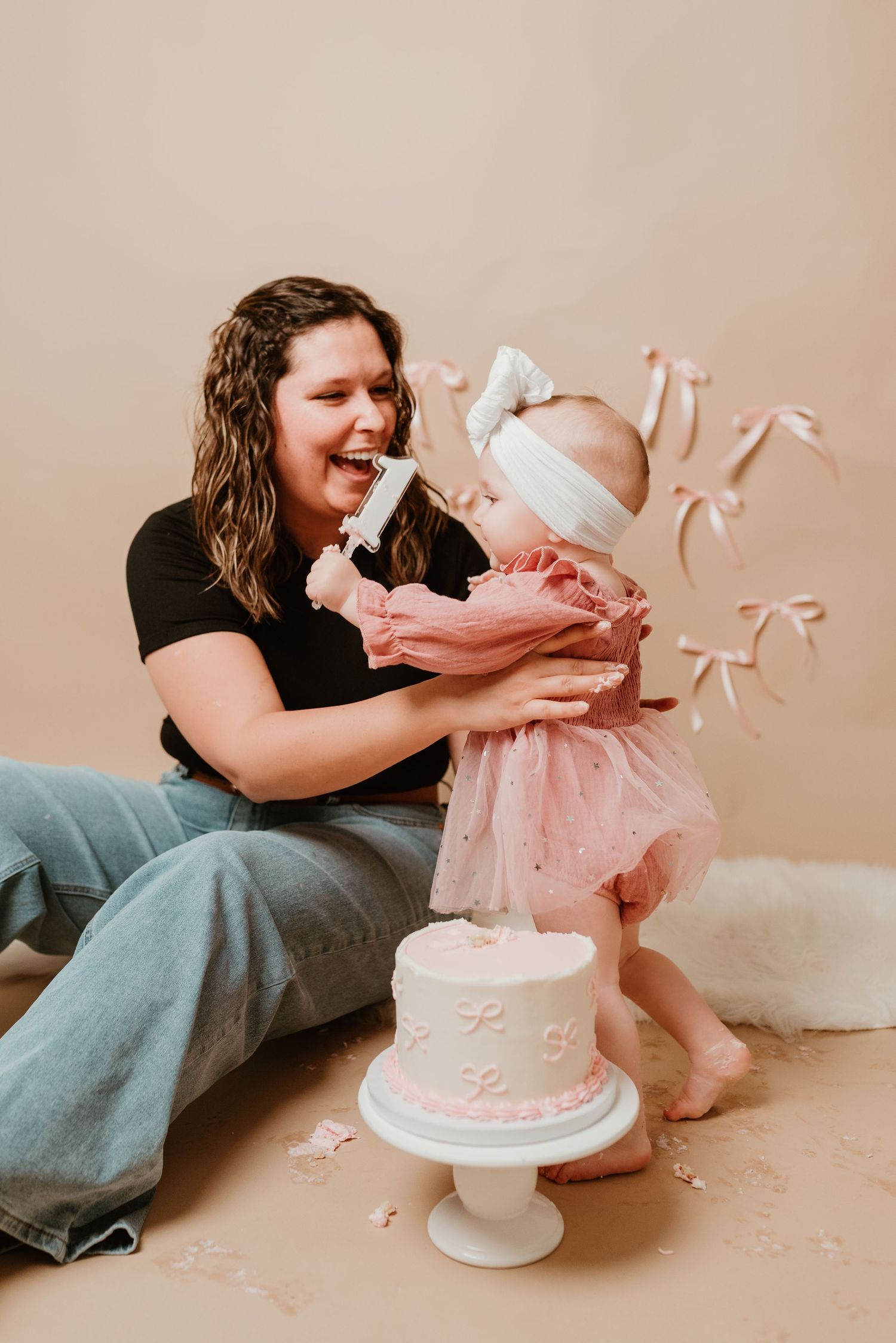 Mother laughs as baby girl in pink tutu stands by first birthday cake smash with pink bow decorations.