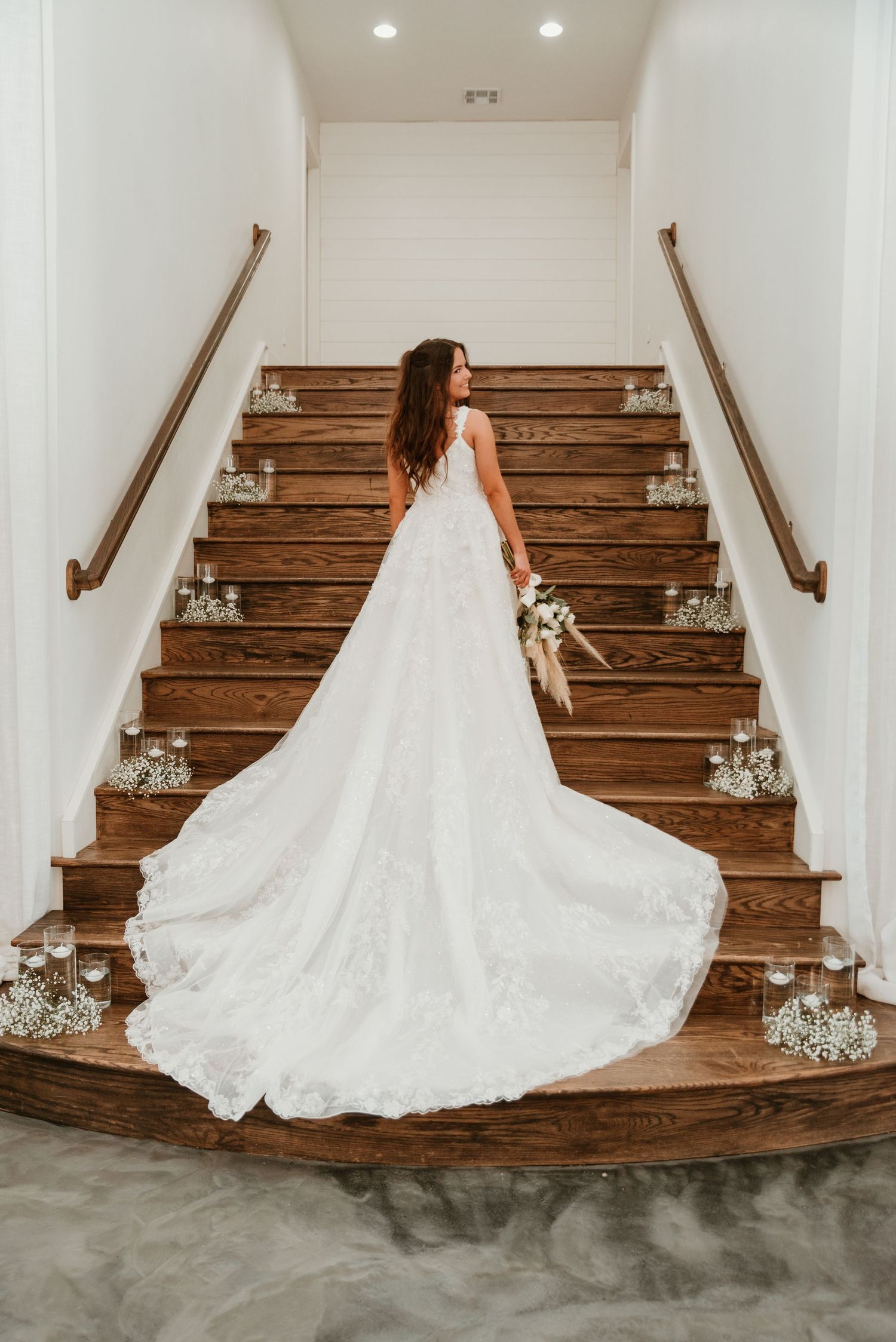 Bride in lace wedding gown with long train poses on wooden staircase decorated with candles and baby's breath.