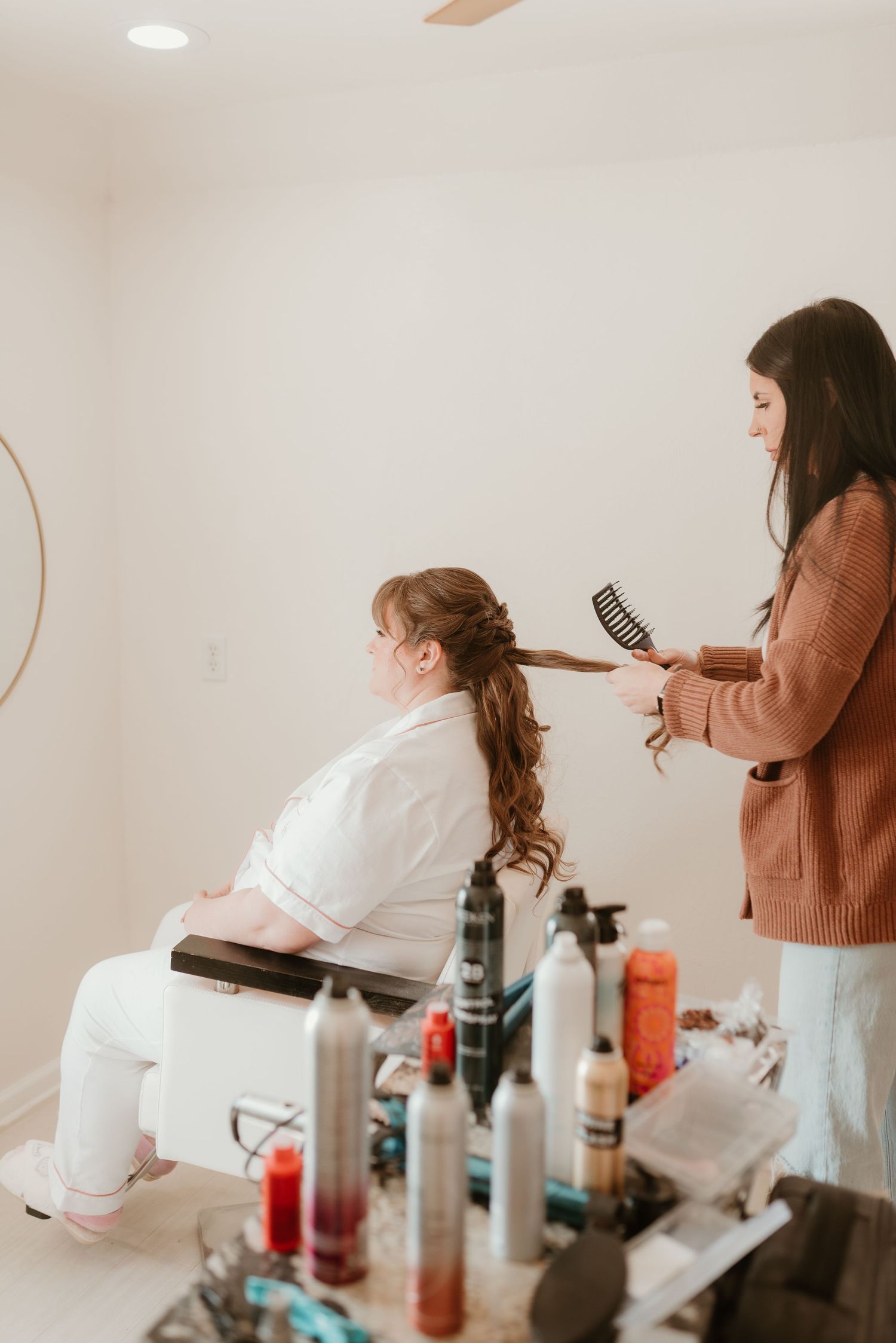 Hair stylist brushing and curling the bride's long hair during wedding day preparations in a bright room.