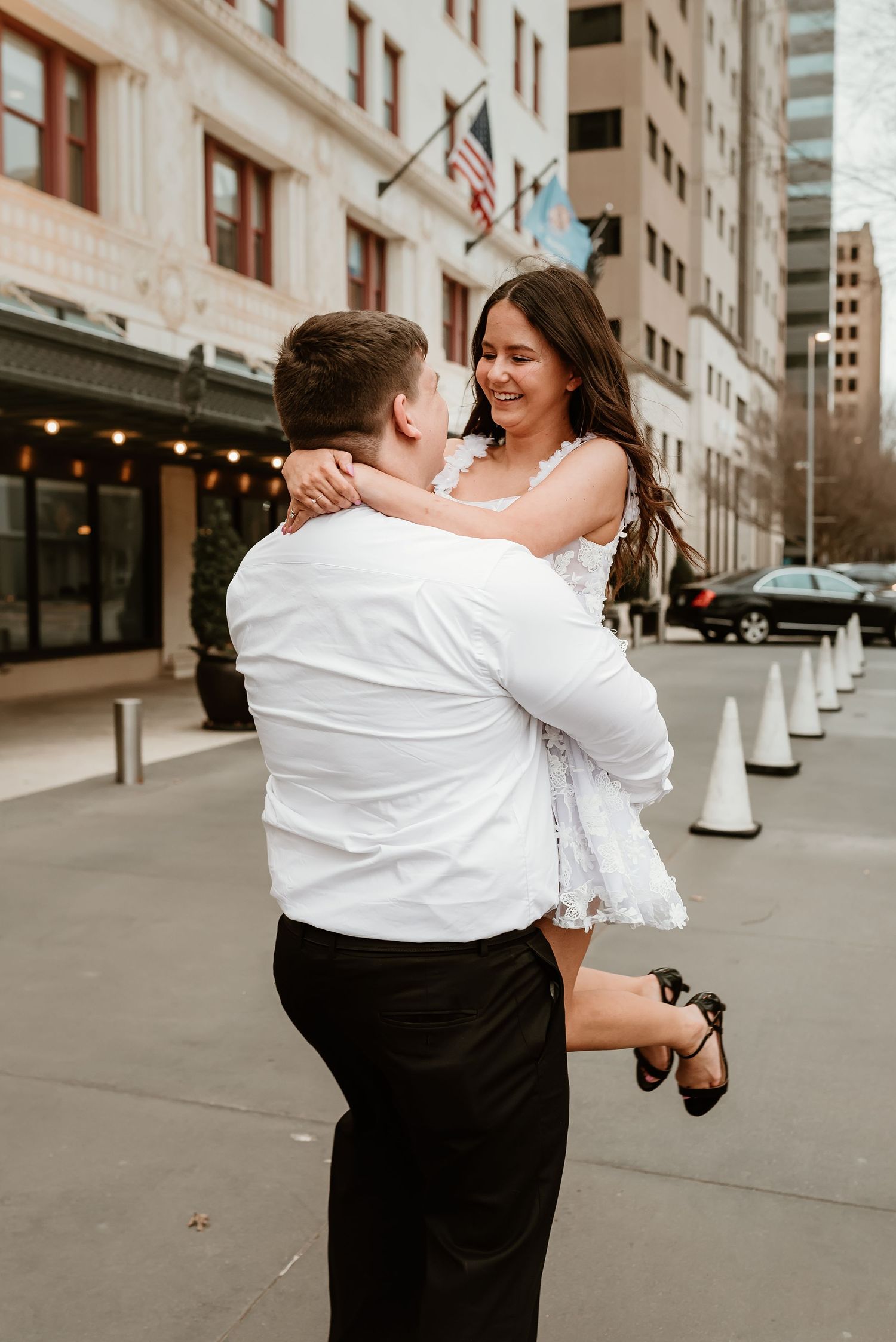 Man lifting smiling woman in white dress on a city street during engagement photos.