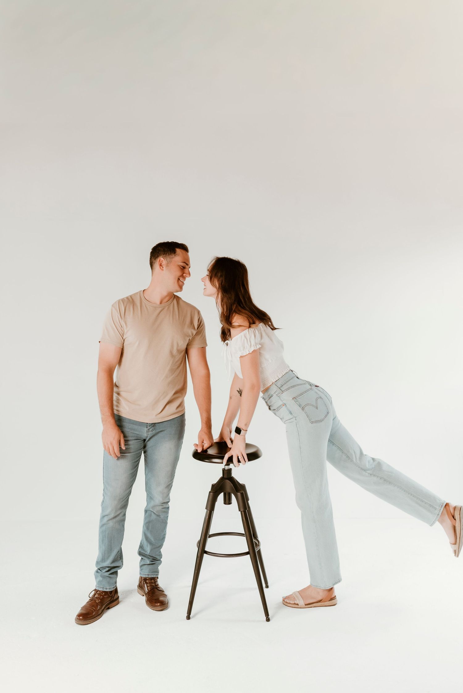 A woman leans on a stool smiling at a man in a bright white studio setting.