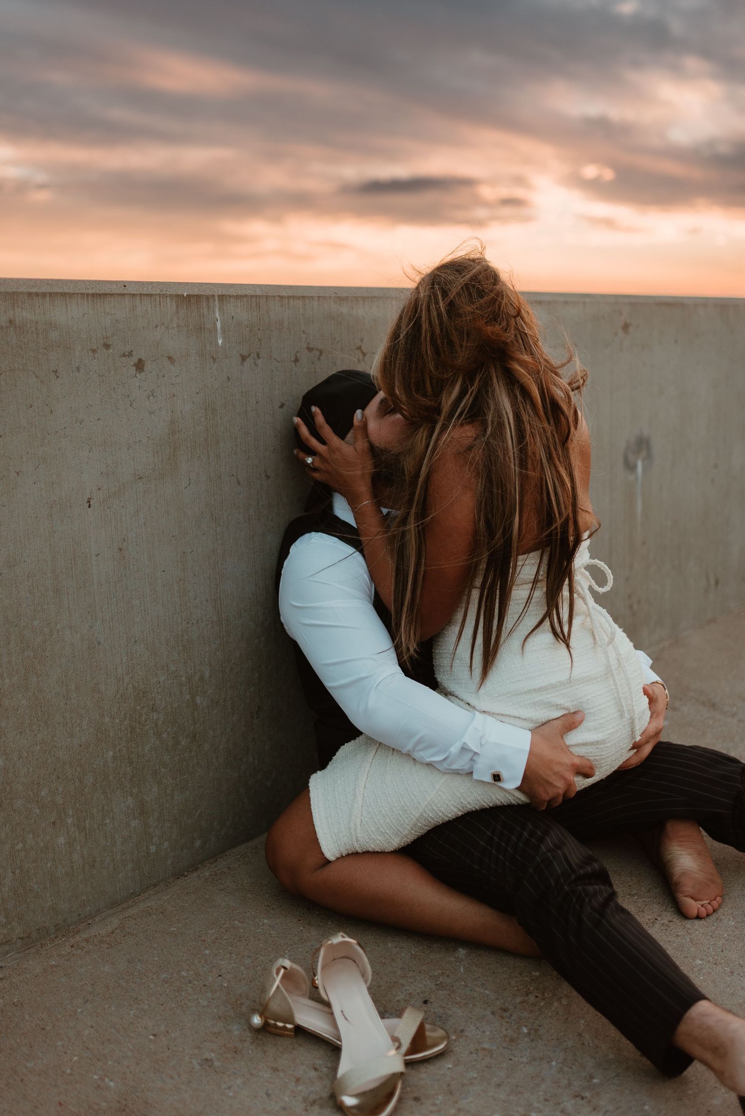 Couple sharing a passionate kiss against a concrete wall at sunset.