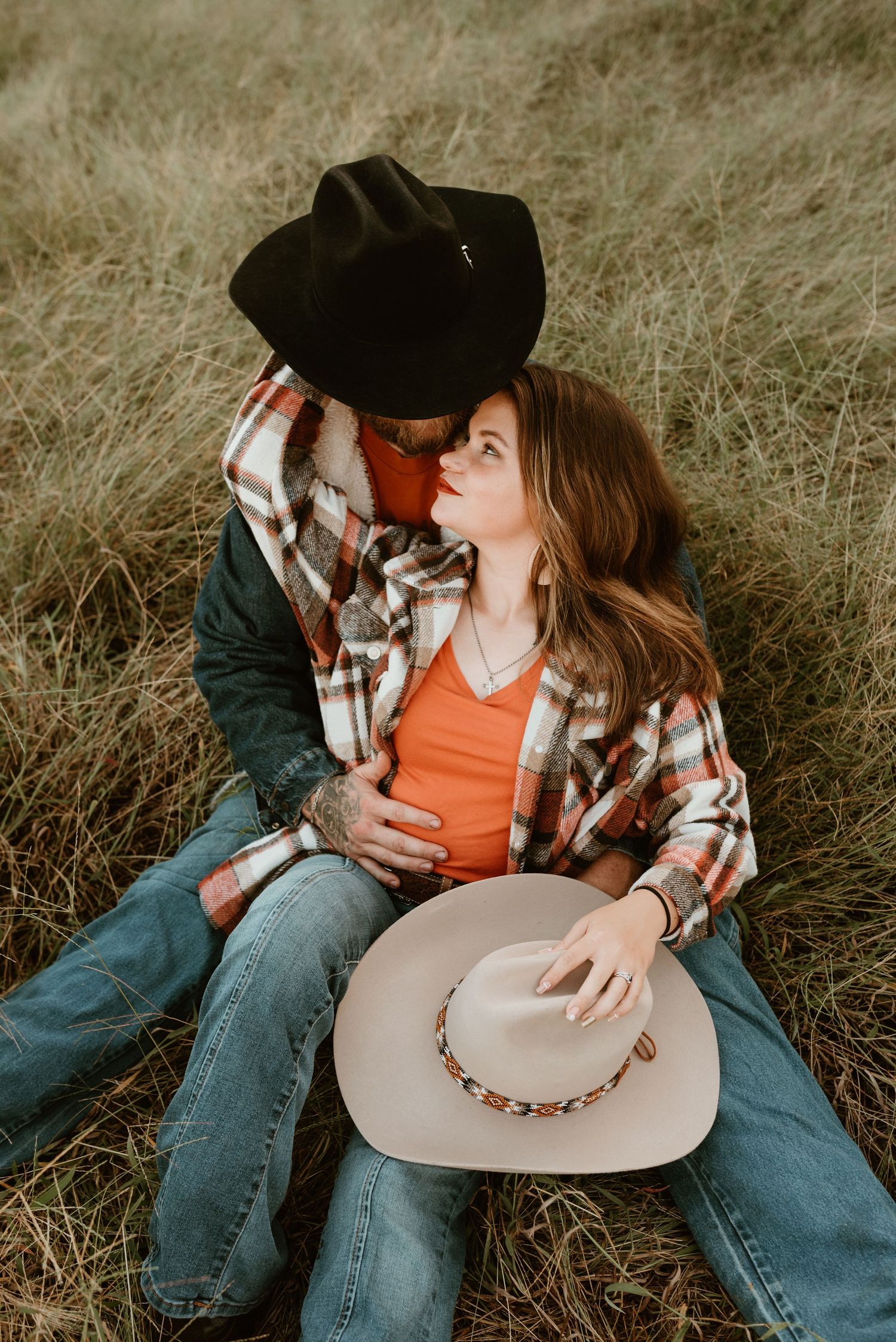 A couple in jeans and plaid sit in a field, sharing a tender moment with cowboy hats.