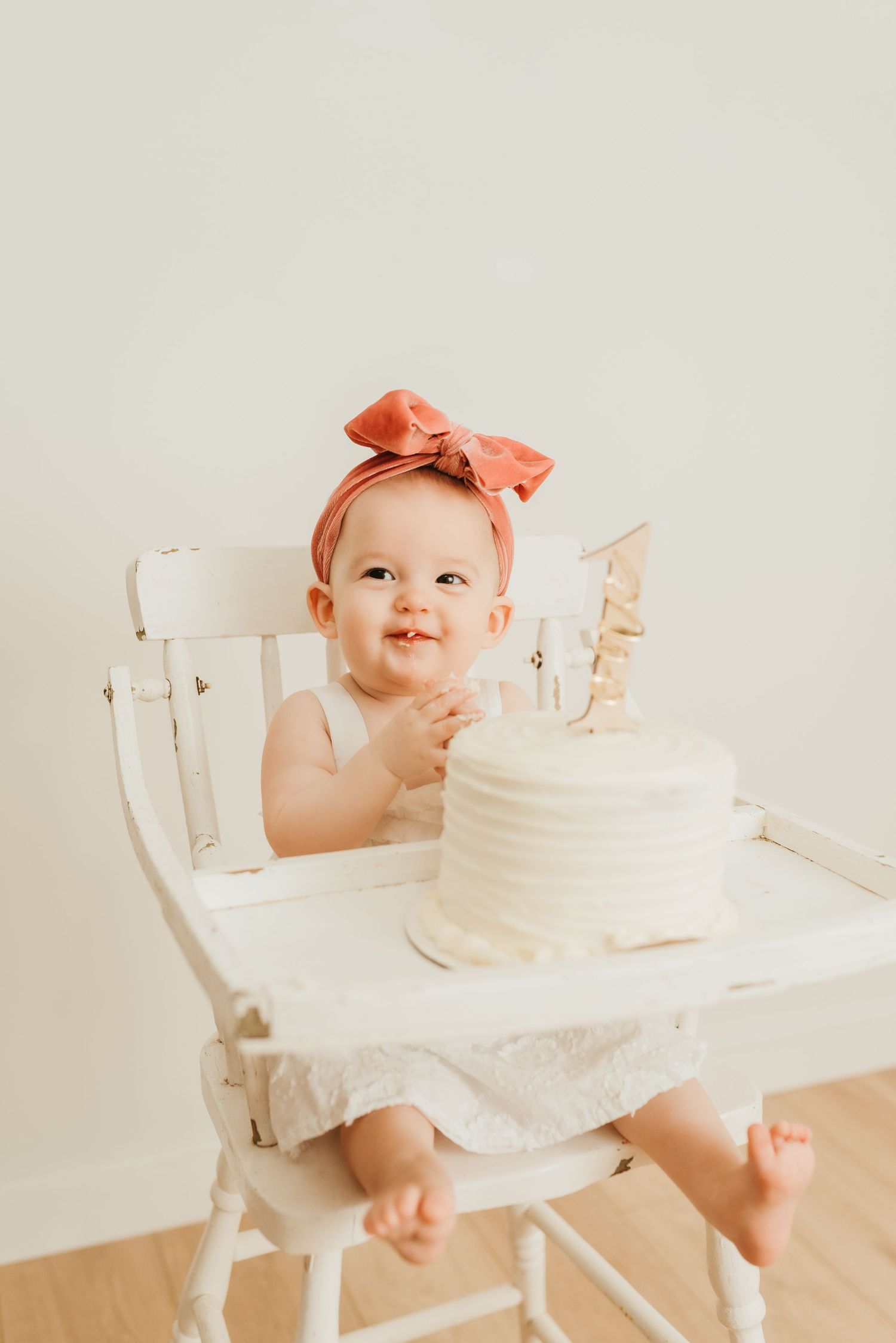 Adorable baby girl in coral bow headband sitting in white high chair with first birthday cake.