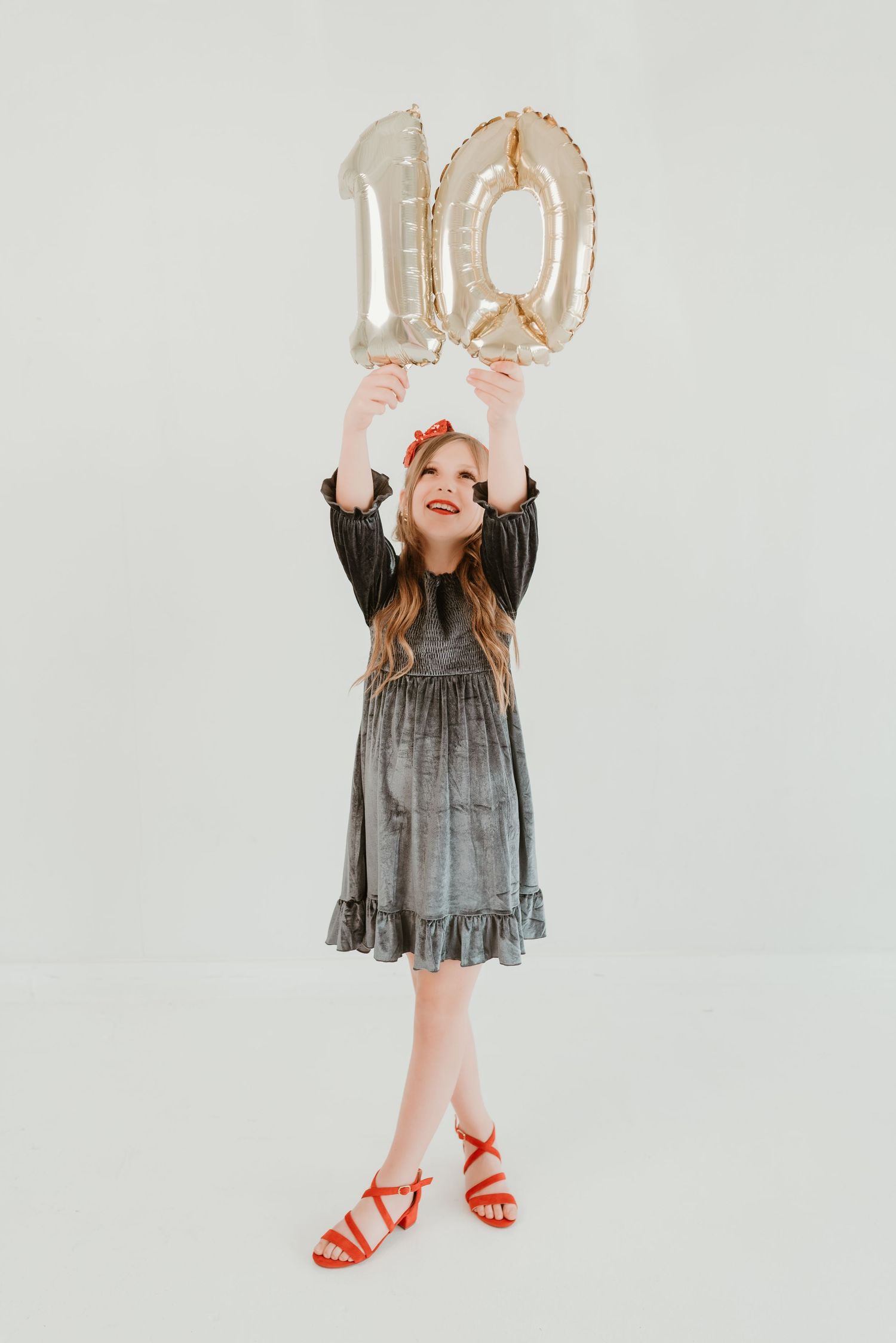 Young girl in gray dress holding gold number 10 balloons overhead on white studio background.