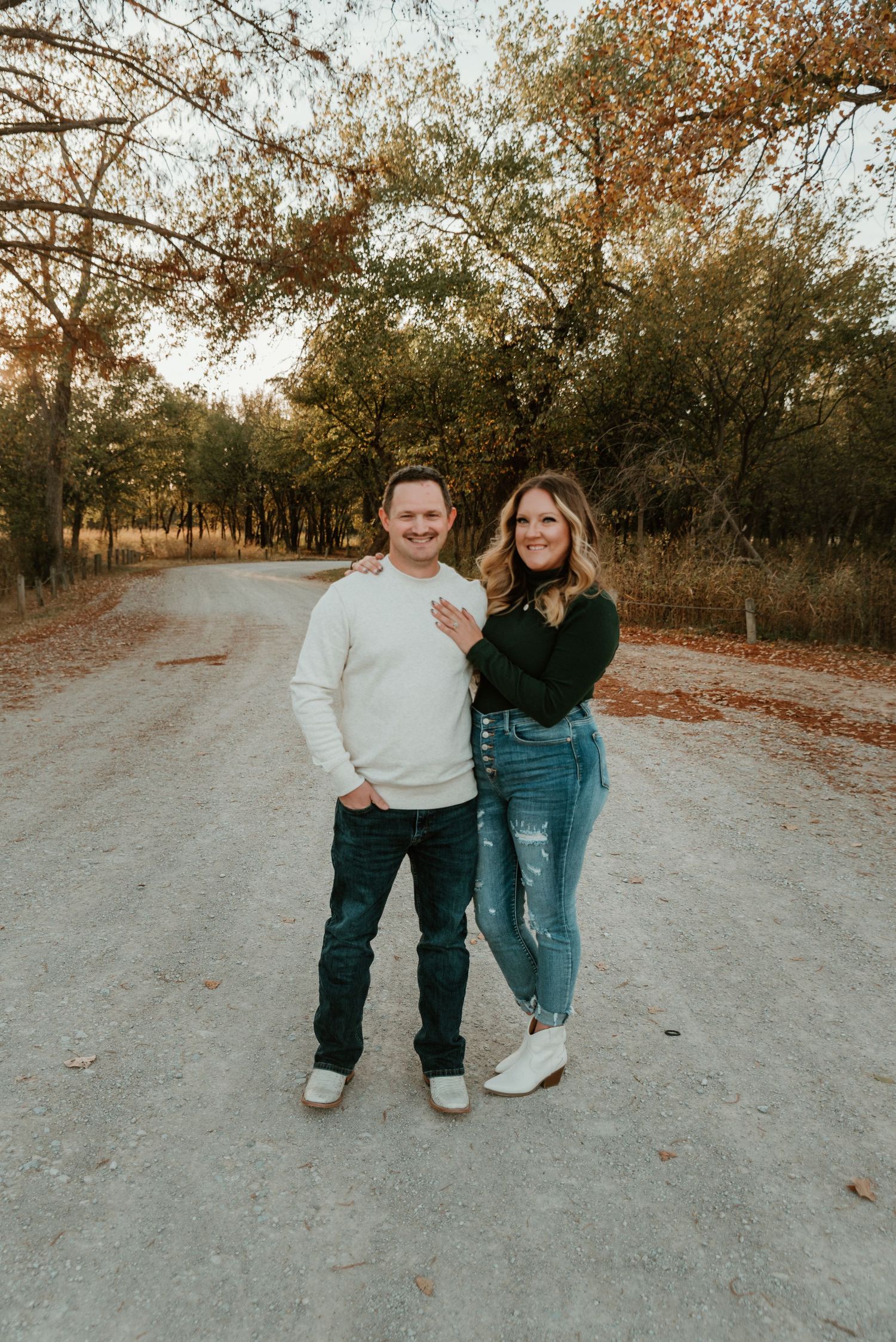 Smiling couple posing together on a dirt path surrounded by autumn trees during an engagement session.