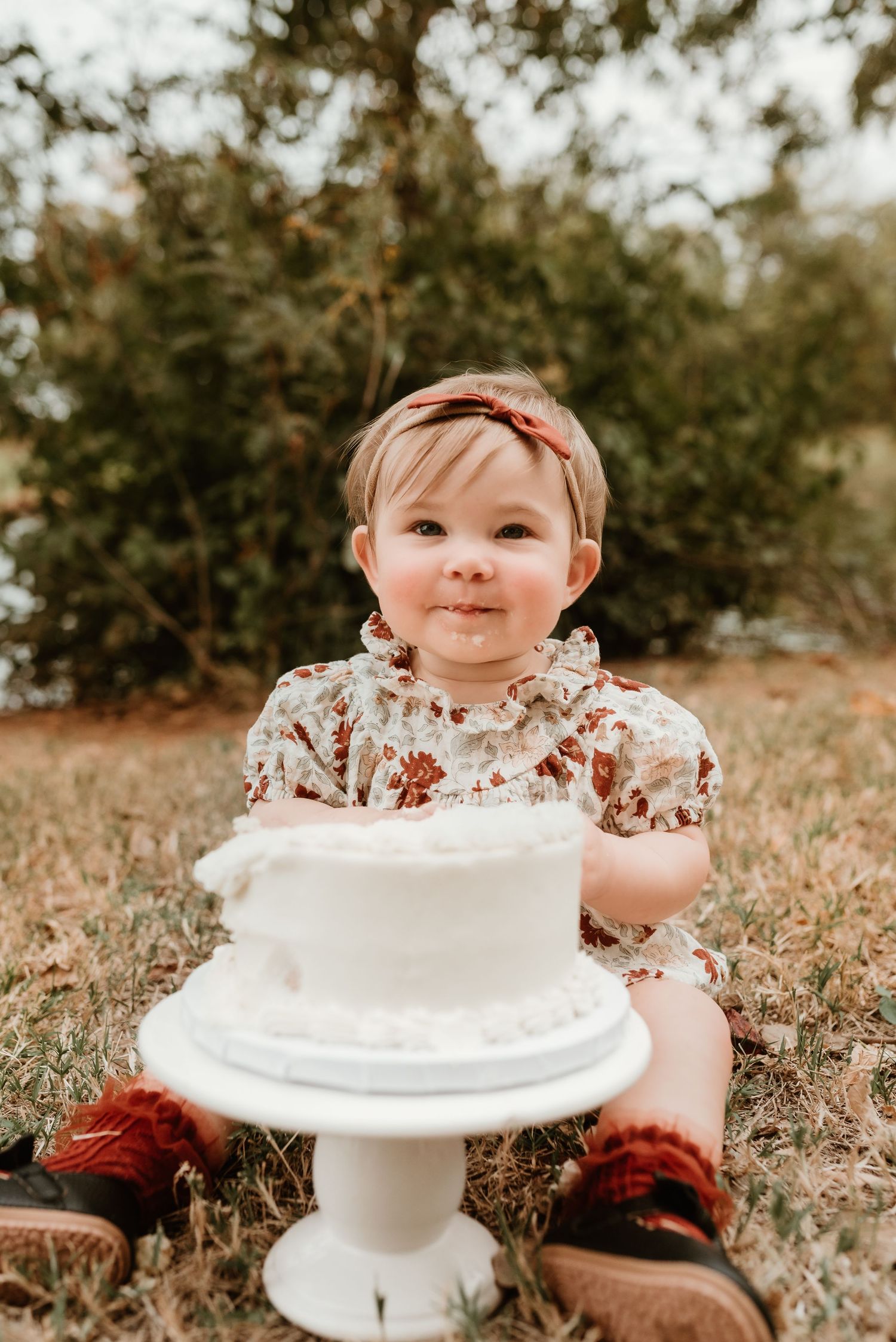 Adorable one-year-old girl in floral outfit sits on grass before a white cake smash cake.