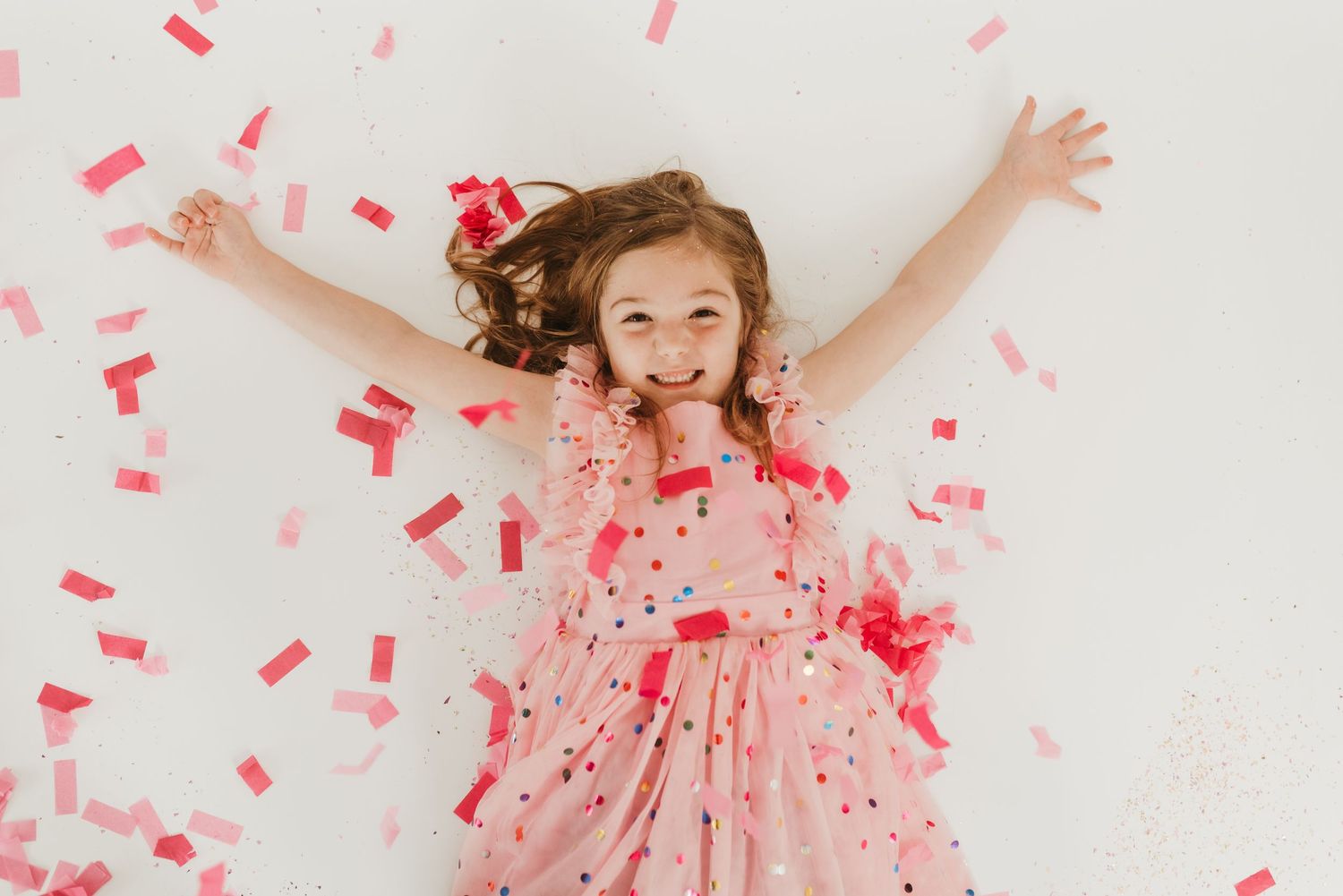 A cheerful young girl in a pink dress lies on the floor surrounded by falling red confetti.
