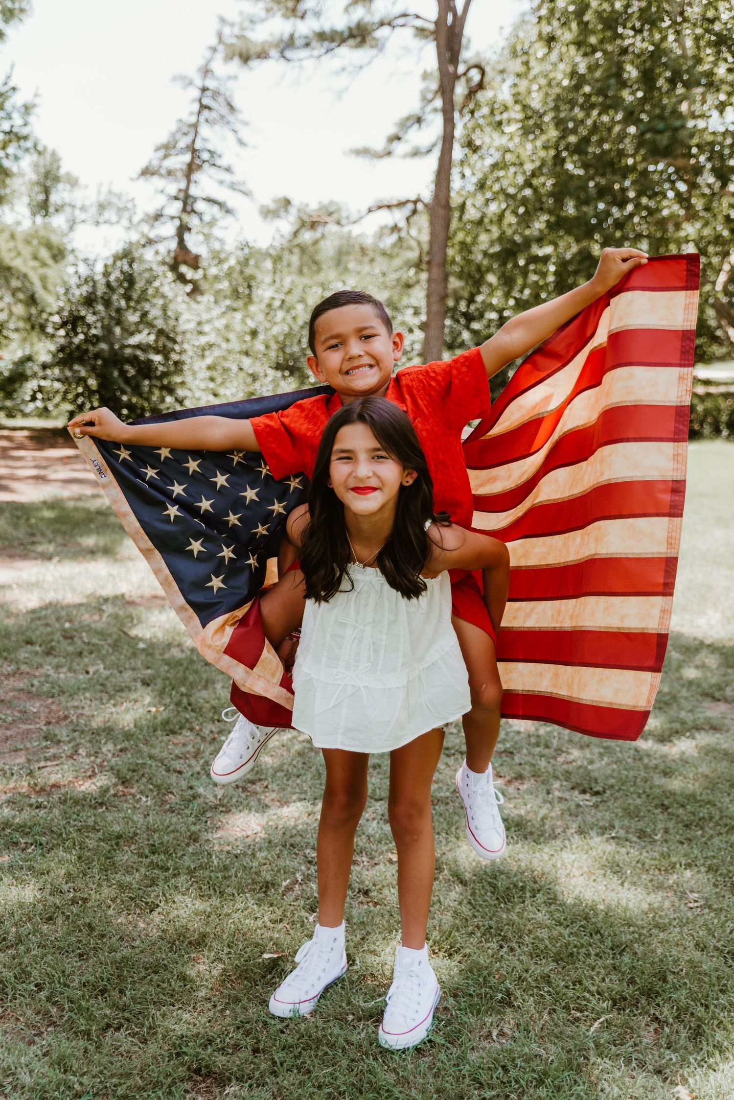 Boy in red shirt and girl in white dress proudly display a large American flag outdoors.