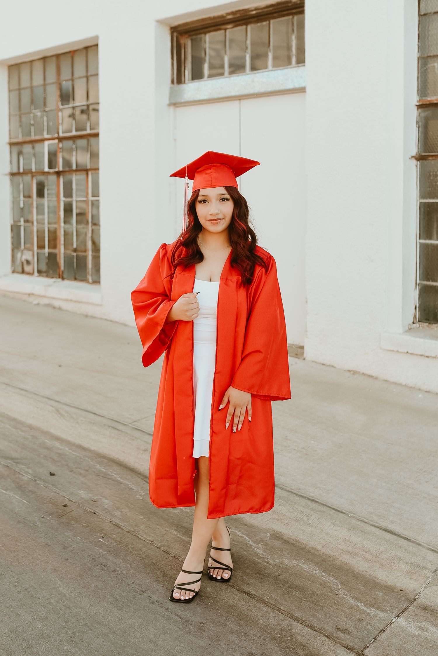 Young woman in red graduation cap and gown smiling outdoors, celebrating academic achievement.