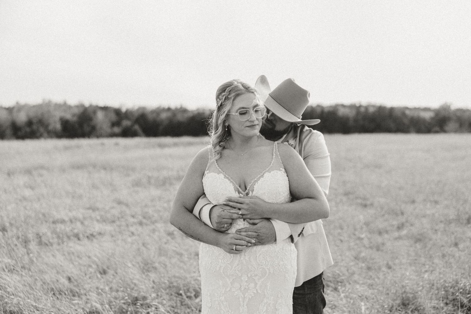 Black and white romantic photo of bride and groom embracing in an open field, groom wearing a cowboy hat.