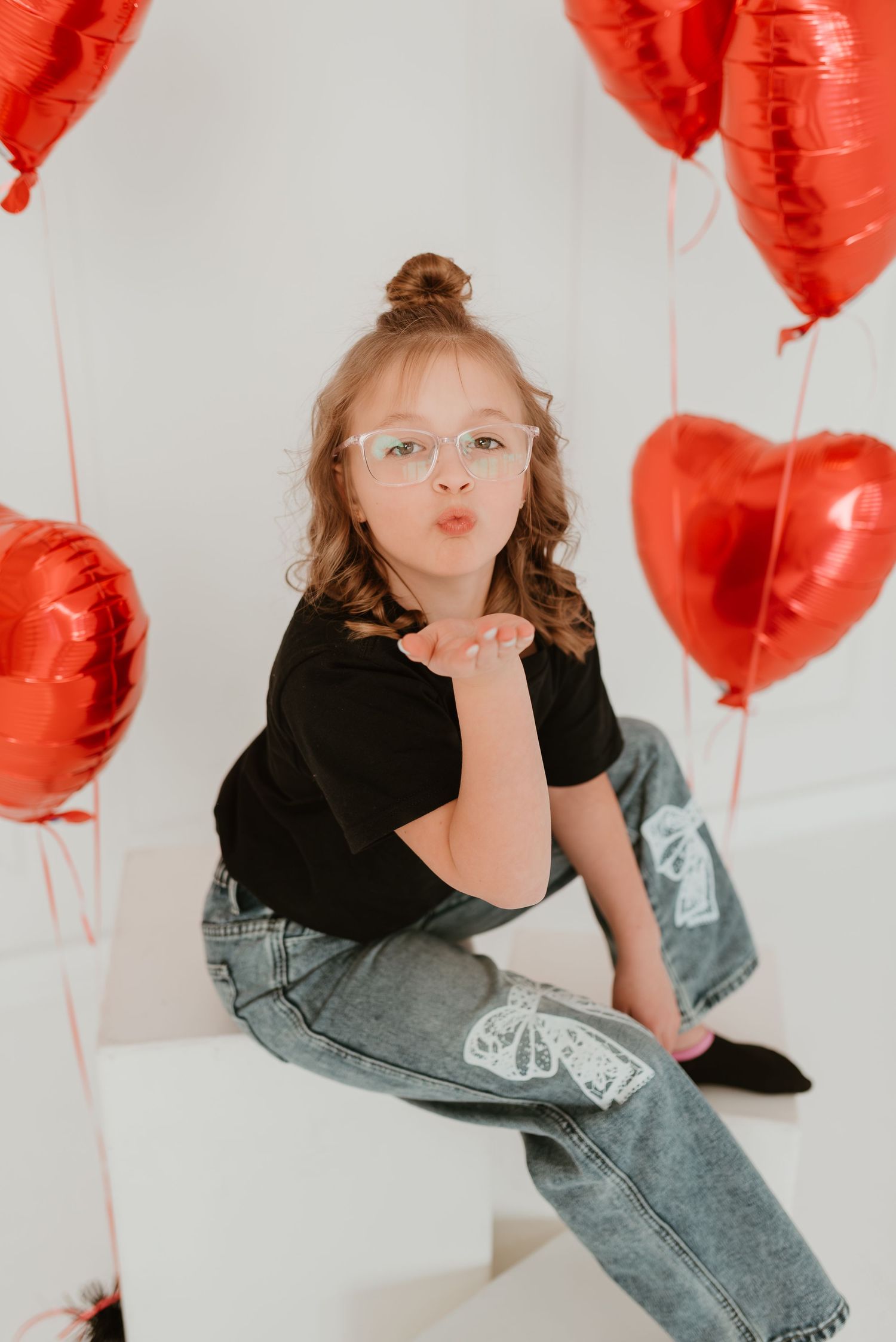 A cute girl with glasses blows a kiss surrounded by red heart-shaped balloons in a bright white studio.