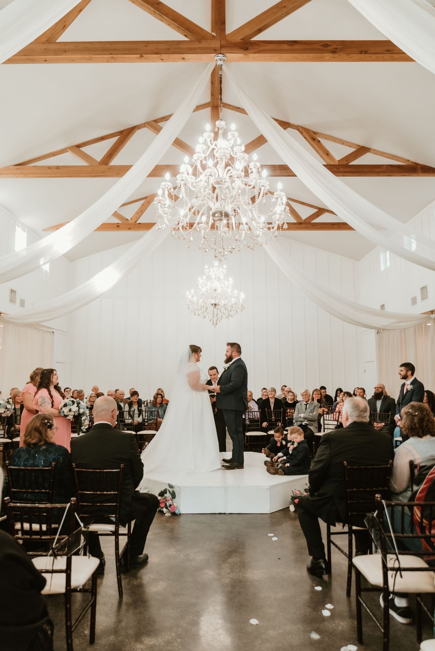 Bride and groom exchanging vows at a stunning barn wedding venue with chandeliers and white draped ceiling.