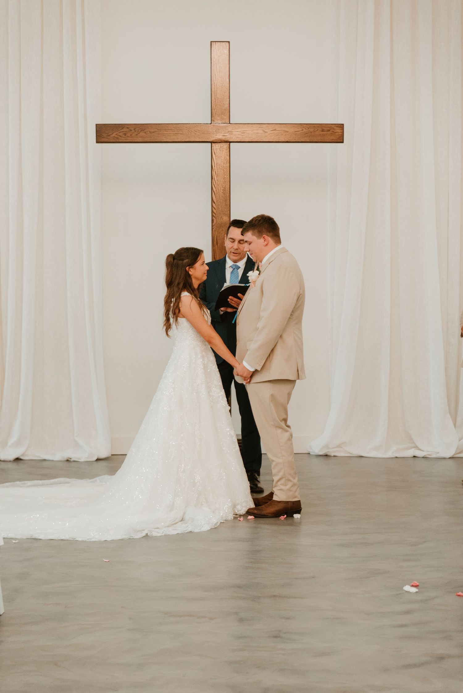 Bride and groom exchange vows before officiant in front of large wooden cross with white drape backdrop.