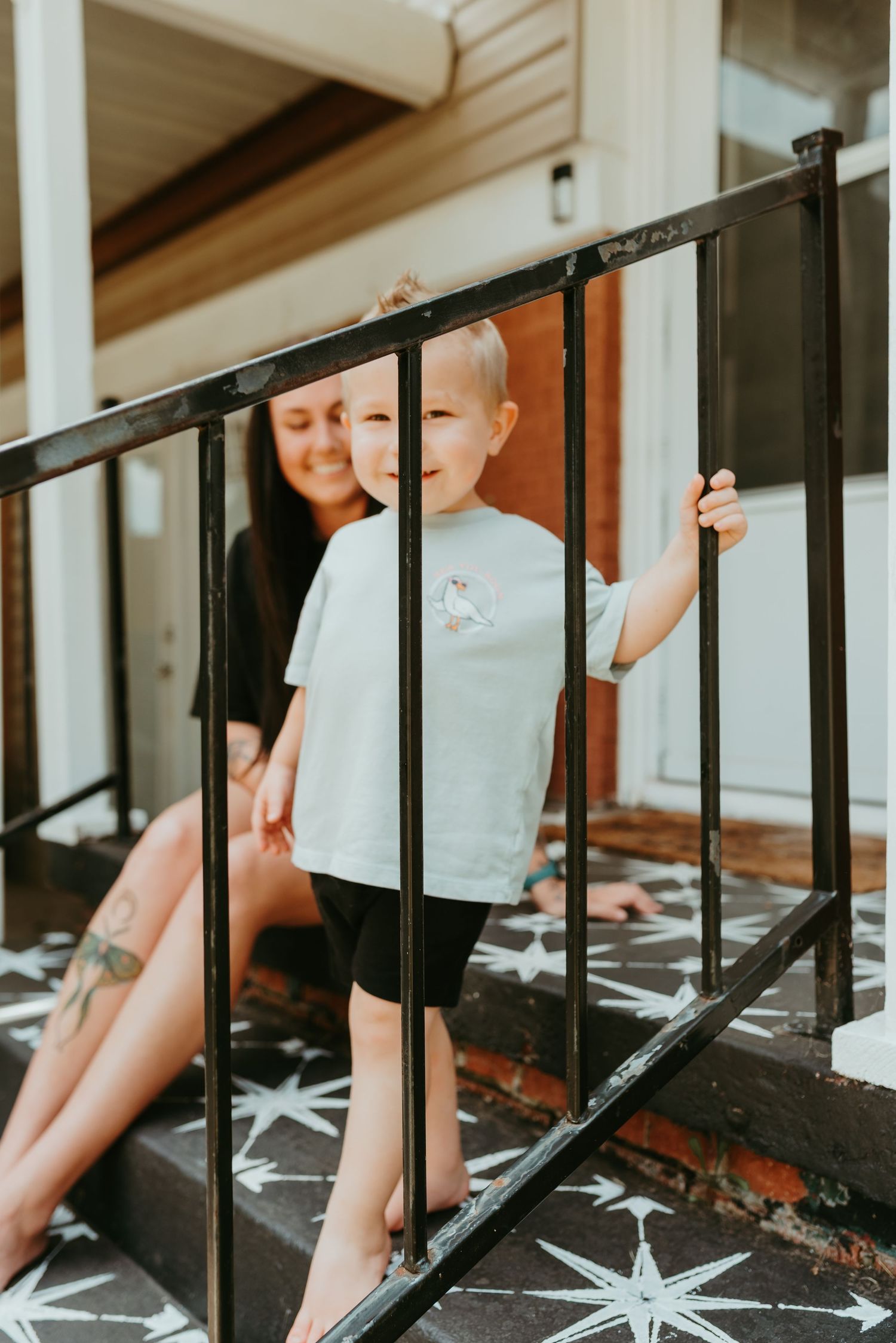 Smiling toddler boy in light tee grips black porch railing while mother sits behind him on decorated steps.