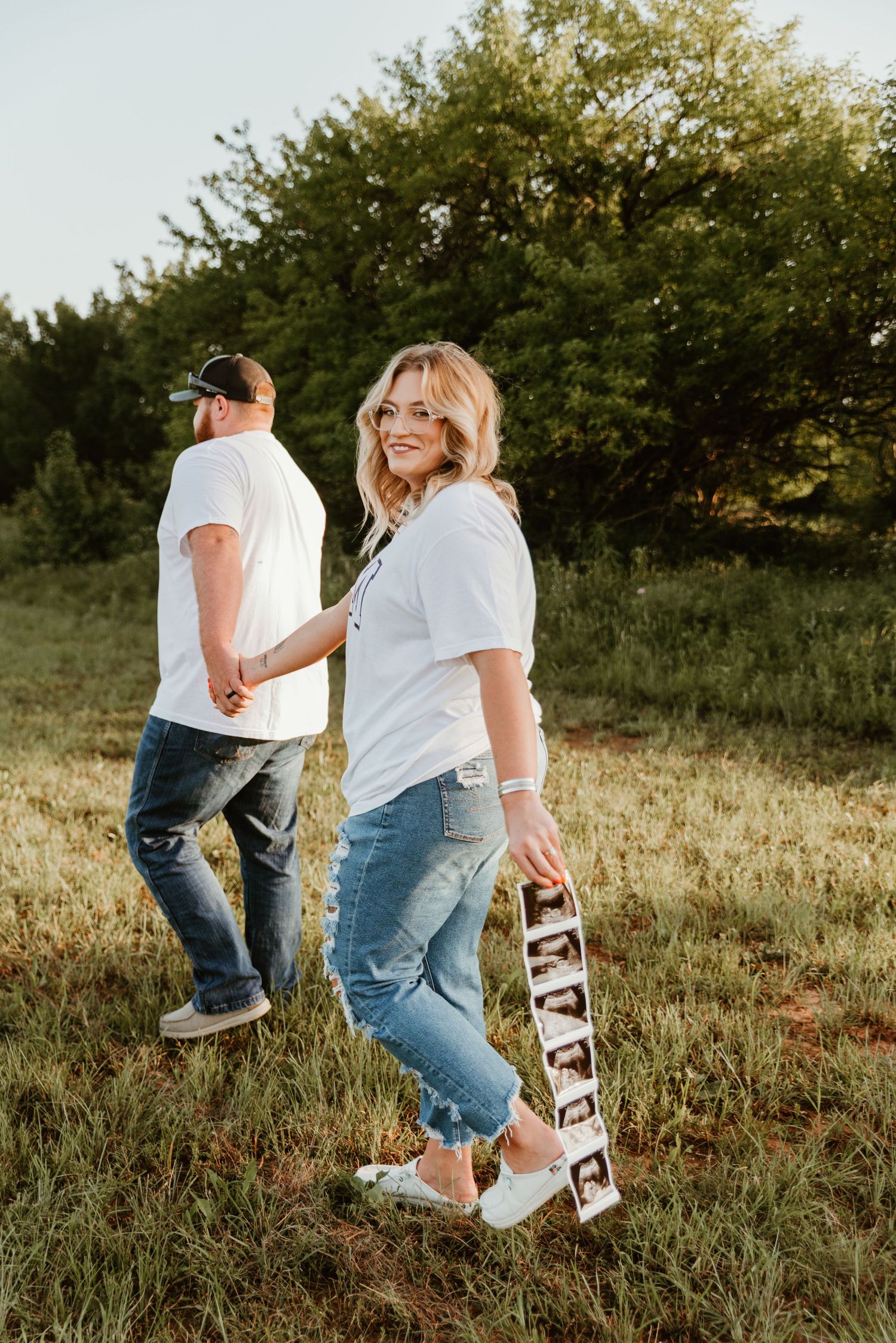 Blonde woman holding ultrasound photos smiles back at camera during outdoor pregnancy announcement.