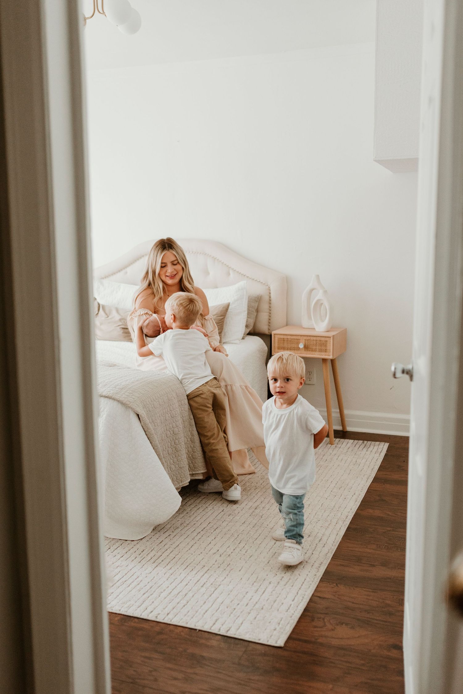 Blonde mother sits on bed smiling while two toddler boys play in bright white bedroom.