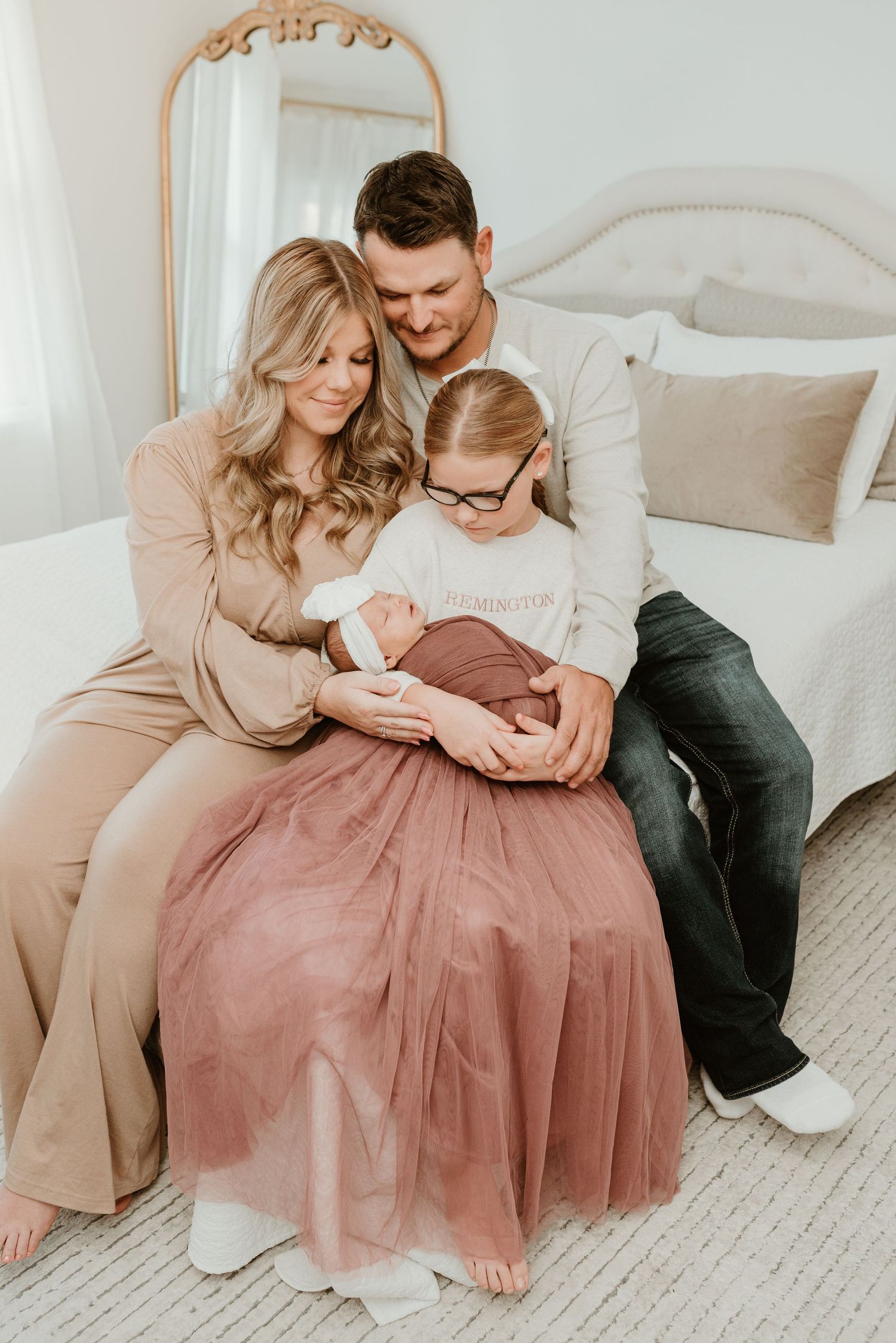 A family of four sits on a bed, parents and young girl in glasses lovingly gazing at their swaddled newborn baby.