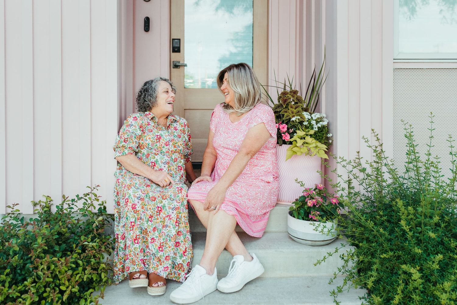 Two women in summer dresses sitting and chatting on a pink front porch surrounded by lush greenery.