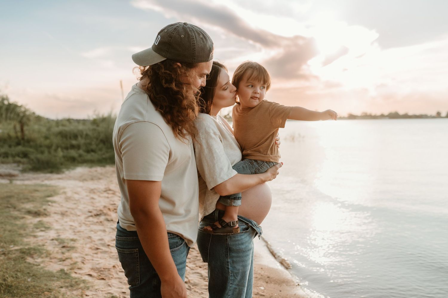 Pregnant mother and father holding toddler on sandy beach at sunset by the water.