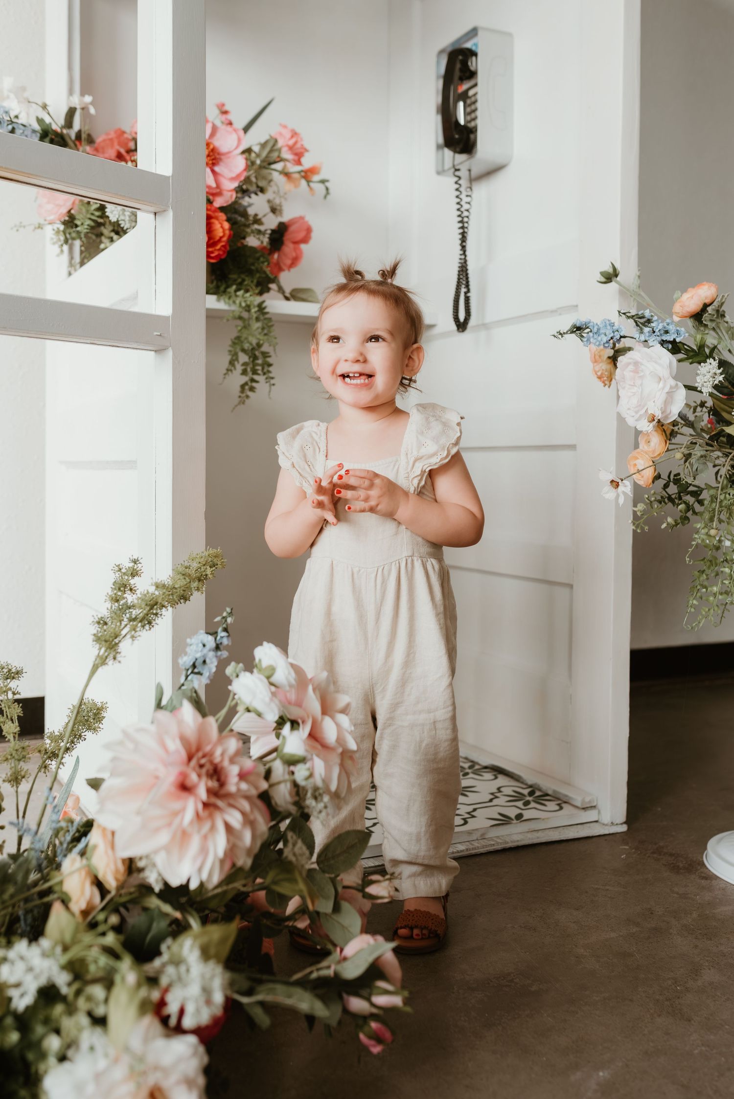 Happy toddler girl in cream romper standing among colorful floral arrangements indoors.