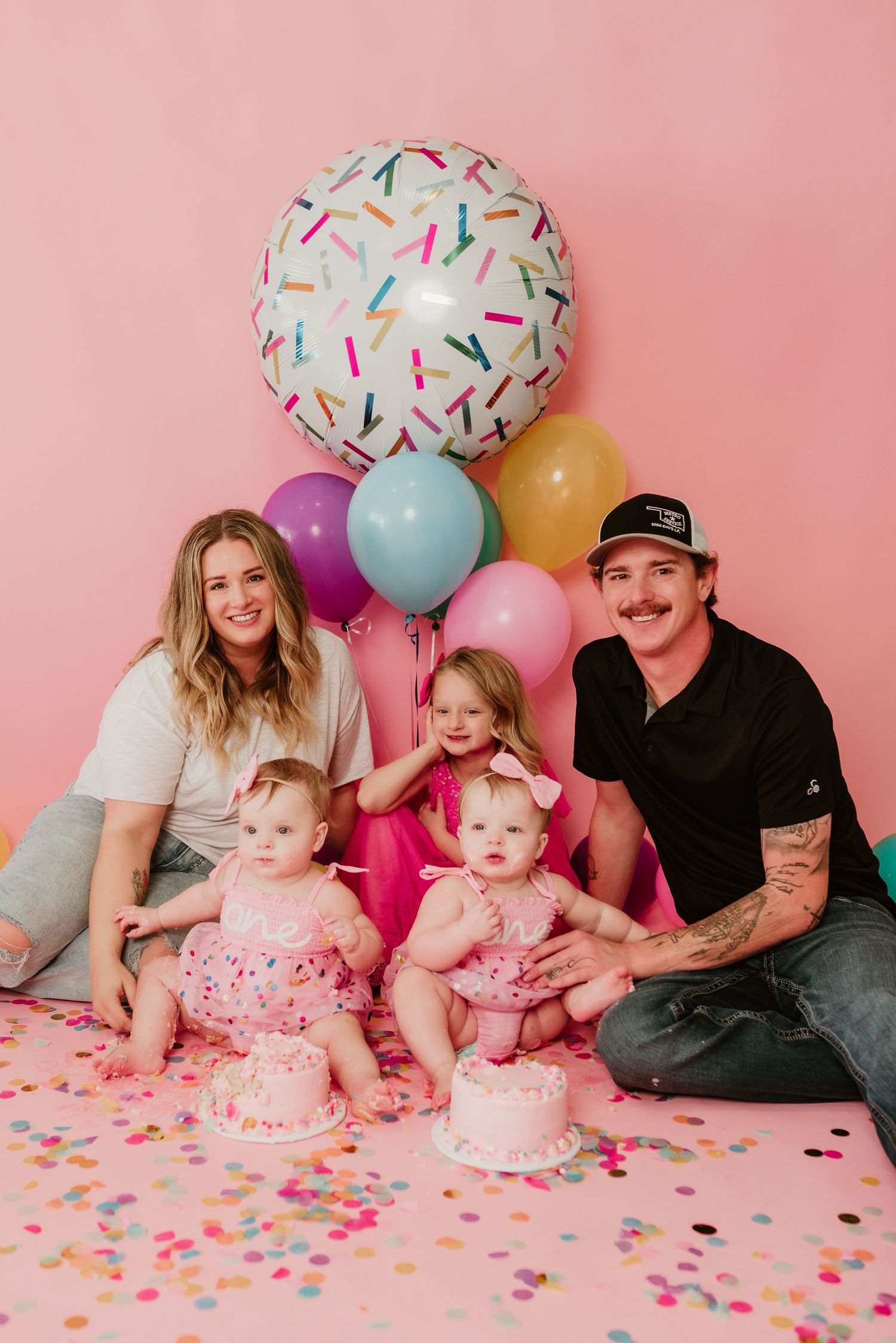 Family of five celebrates twin girls first birthday with colorful balloons and smash cakes on pink backdrop.
