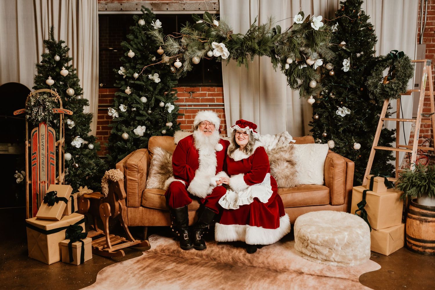 Santa and Mrs. Claus pose on a leather sofa surrounded by elegant white Christmas tree decorations.
