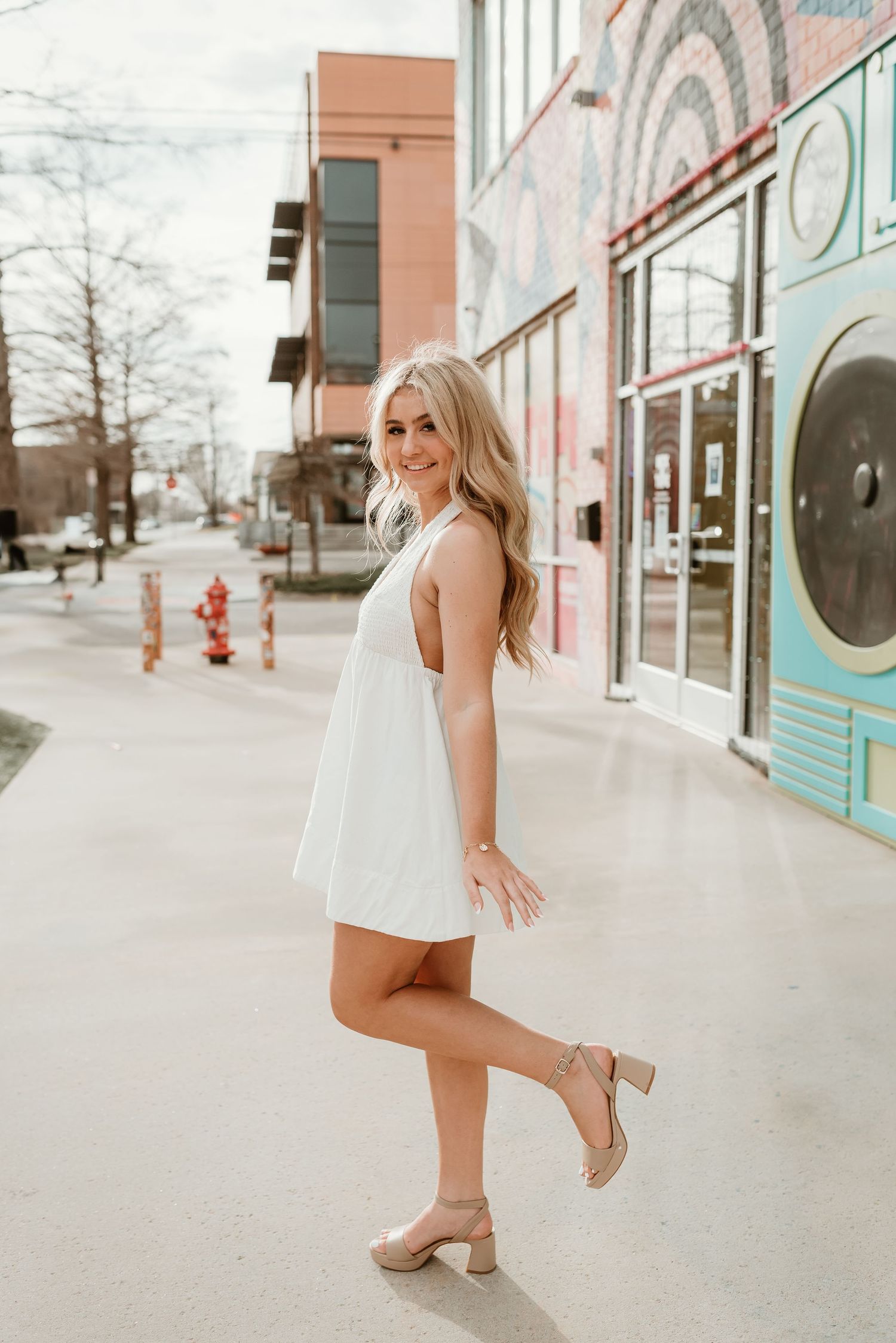 A blonde woman in a white mini dress poses on an urban sidewalk with colorful murals.
