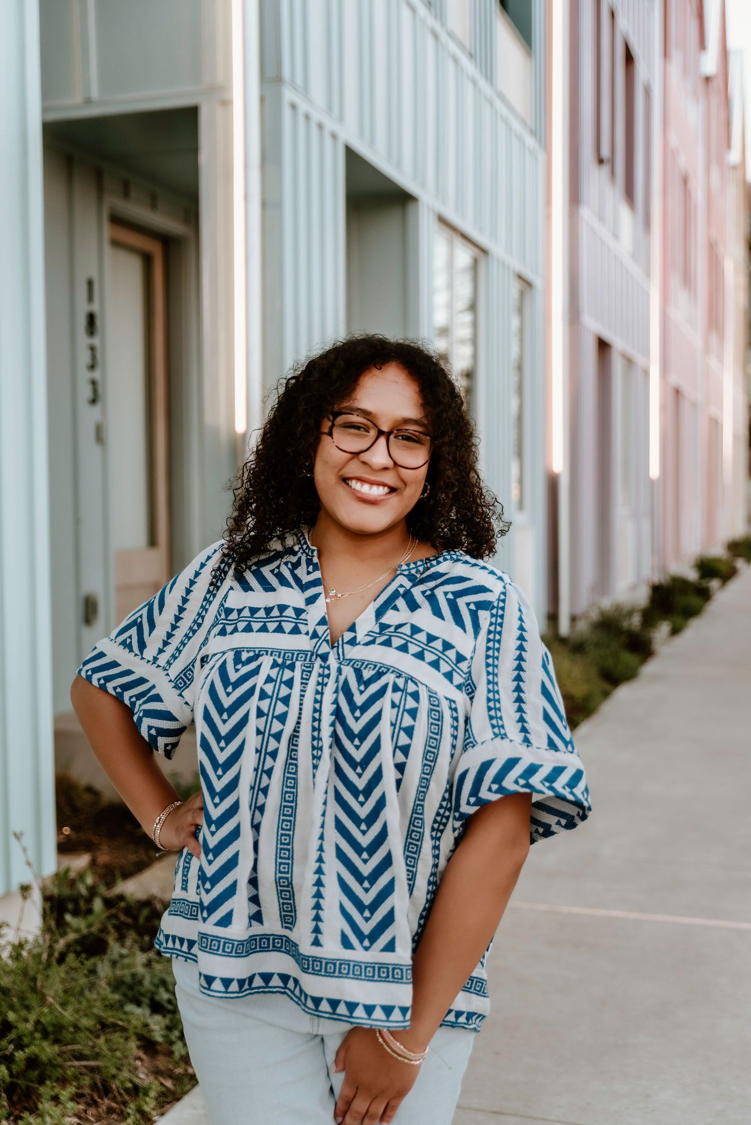 Smiling woman in blue patterned top and glasses posing on urban sidewalk between colorful buildings.