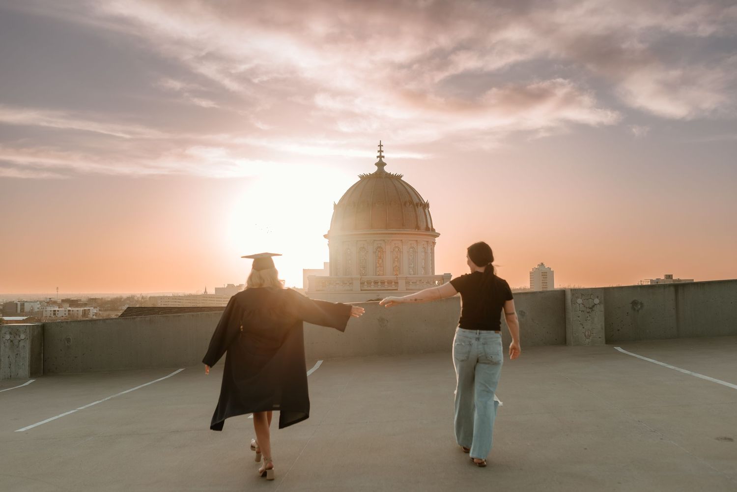 Two women walk hand in hand on a rooftop at sunset near a domed building.