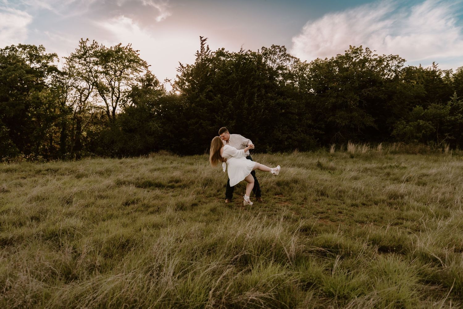 A man lifts and spins his partner in a white dress through a lush green meadow at golden hour.
