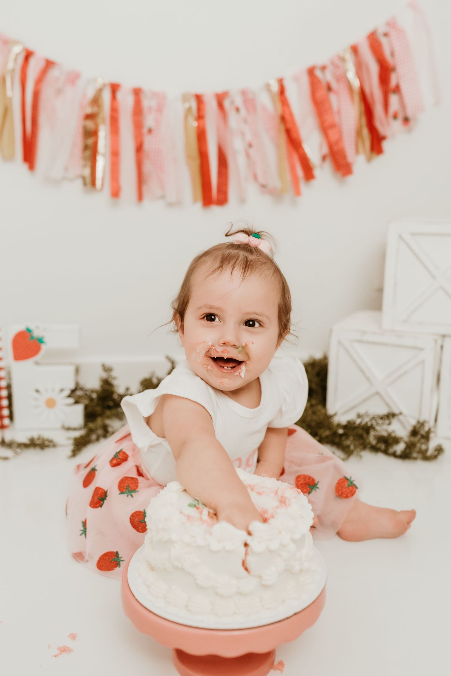 Smiling baby girl in strawberry outfit smashing white birthday cake with frosting on her face.