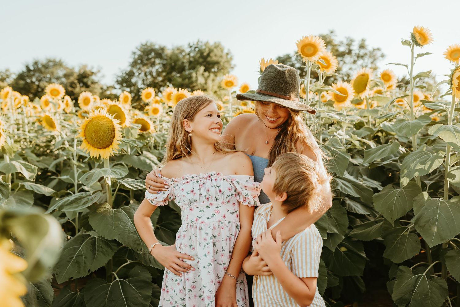 A mother in a hat embraces her two daughters while standing in a sunny sunflower field.