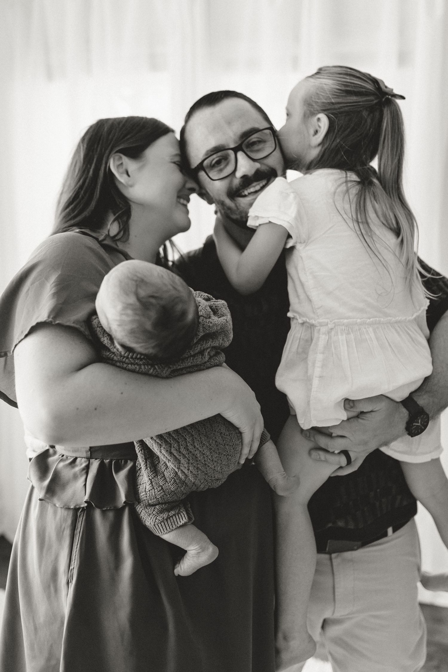 Black and white photo of smiling family with dad holding toddler girl and mom cradling newborn baby.