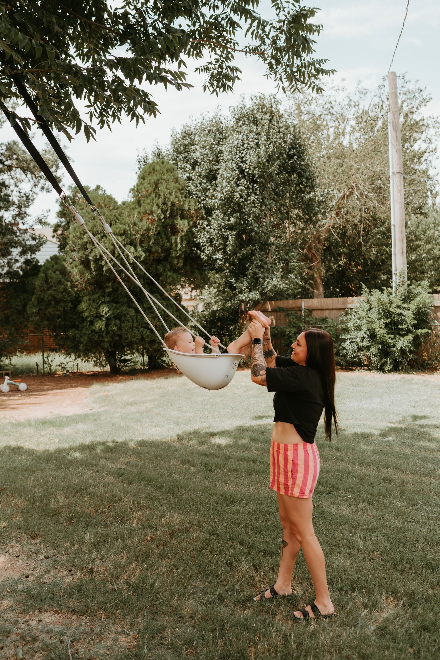 Young mother in black top and striped shorts pushes baby in hanging outdoor hammock swing in green backyard.