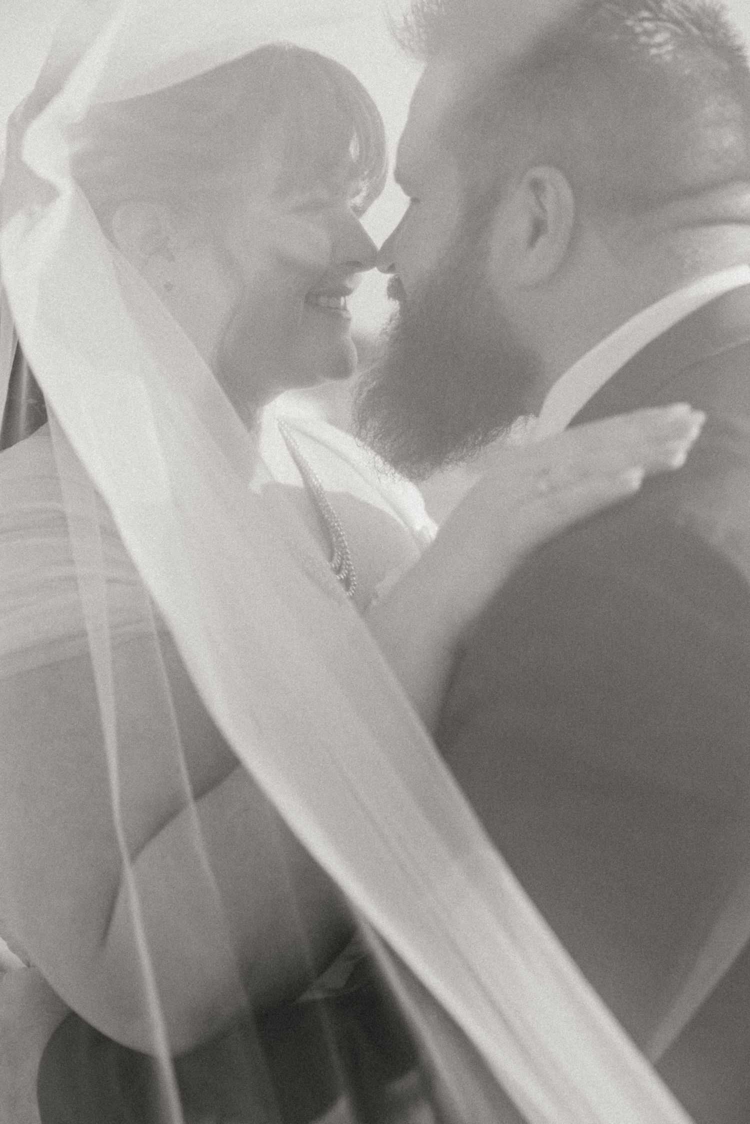 Romantic black and white photo of bride and groom smiling nose to nose under a sheer bridal veil.