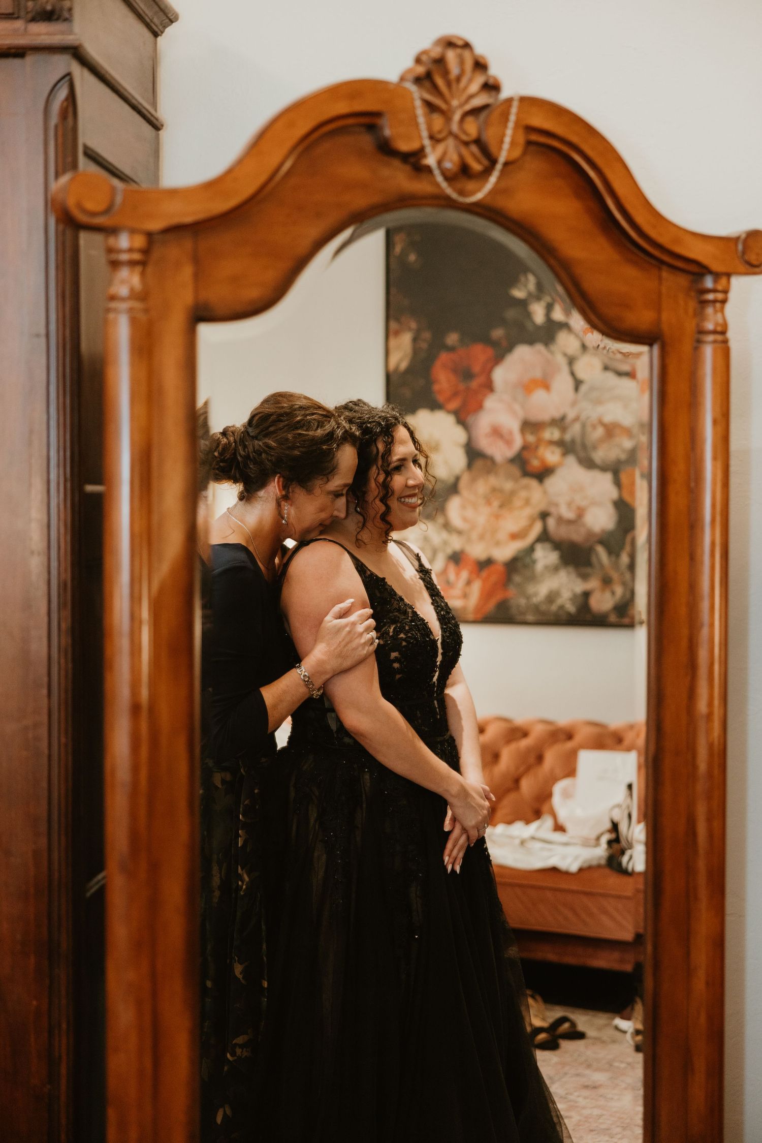 Bride in black wedding gown shares a tender moment reflected in ornate wooden mirror before ceremony.
