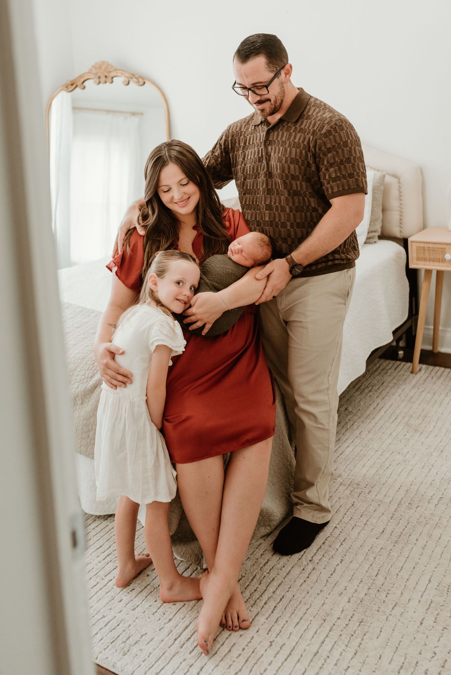 A family of four poses in a bright bedroom, mother in red dress and father in brown shirt holding their newborn and toddler.