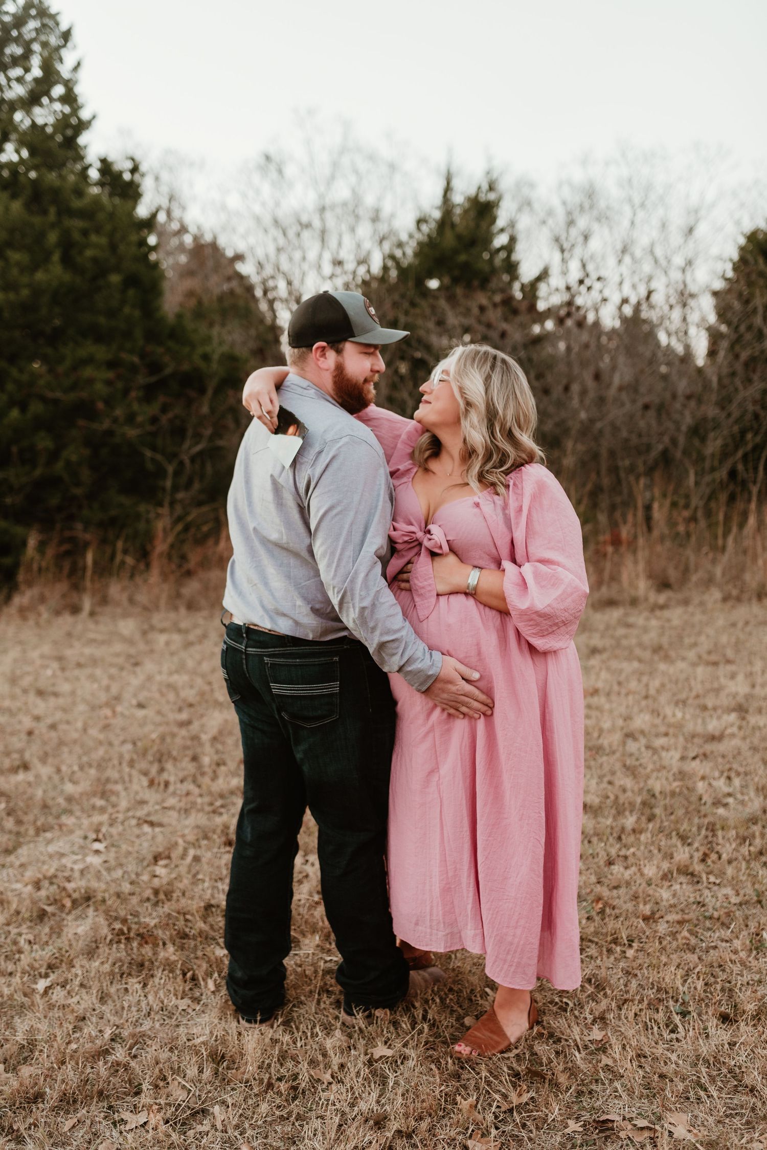 A pregnant woman in a pink maternity dress shares a romantic moment with her partner in an outdoor field setting.