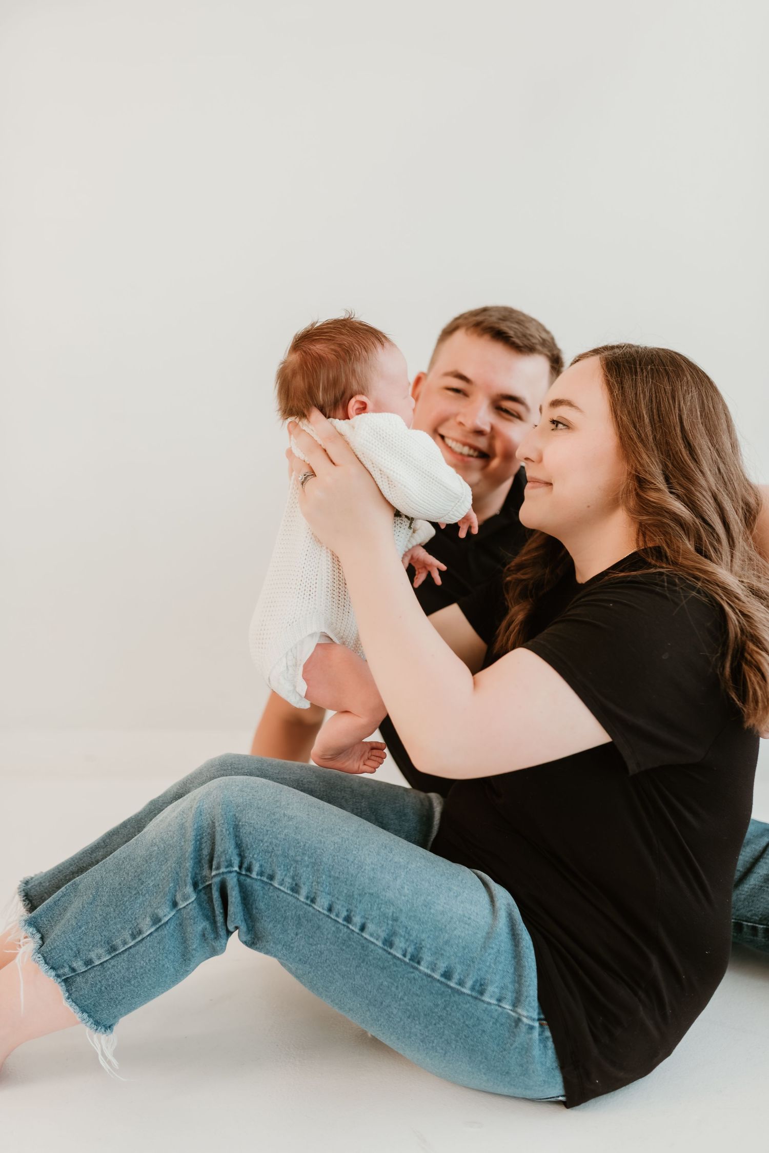 A smiling couple sits on a white studio floor lovingly holding their newborn baby between them.