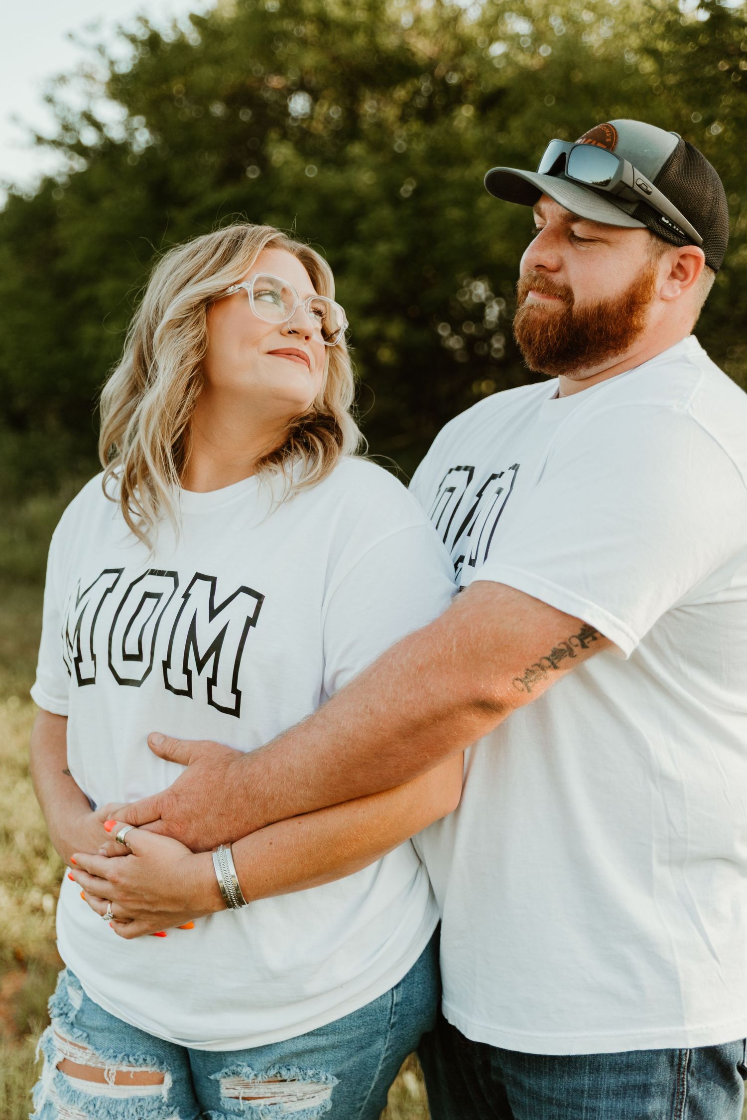 Man embracing pregnant woman from behind outdoors, both wearing matching MOM and DAD shirts.