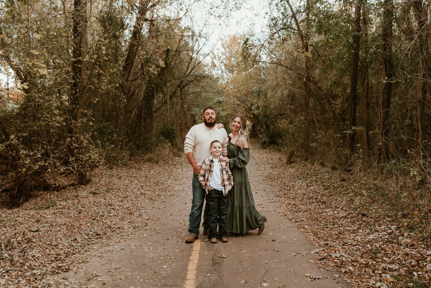 Family of three poses on a tree-lined road during autumn outdoor photo session.