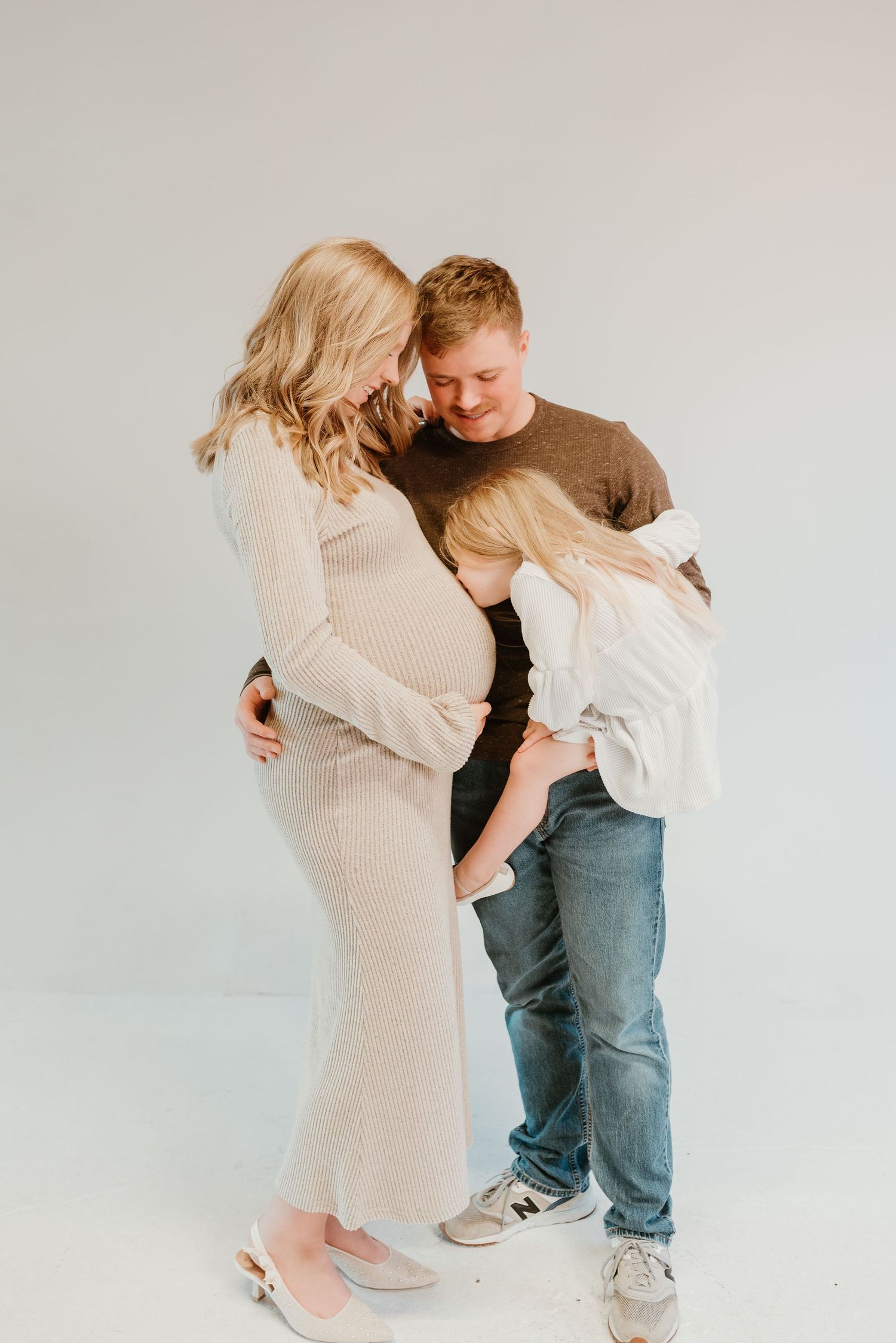 Pregnant blonde woman in cream dress embraced by husband and child in bright studio.