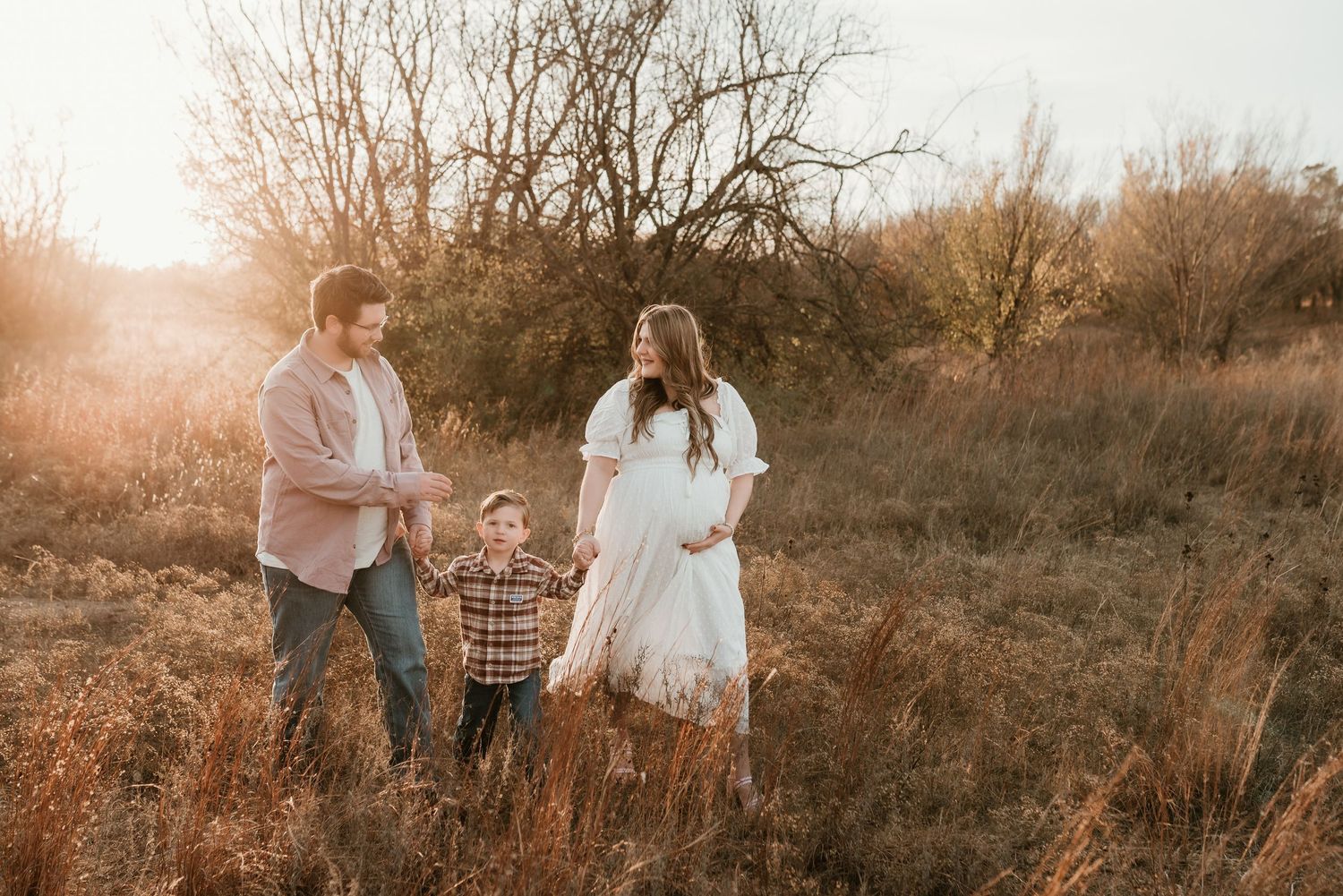 Expecting family with toddler son walks through golden field during sunset maternity session.