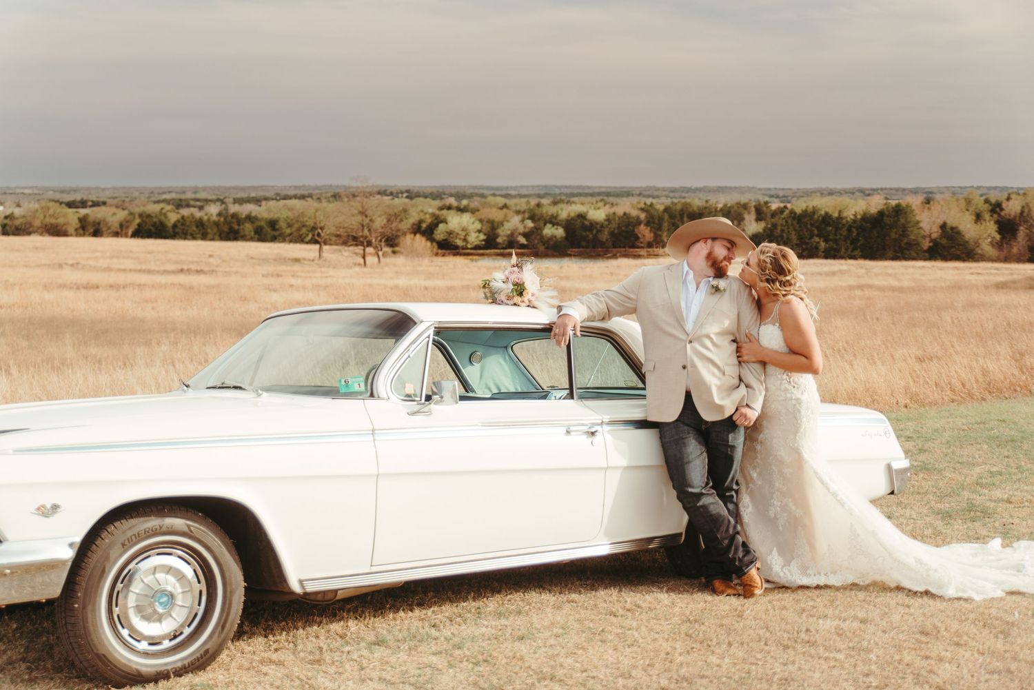 Bride and groom share a romantic moment leaning against a vintage white classic car in an open field.