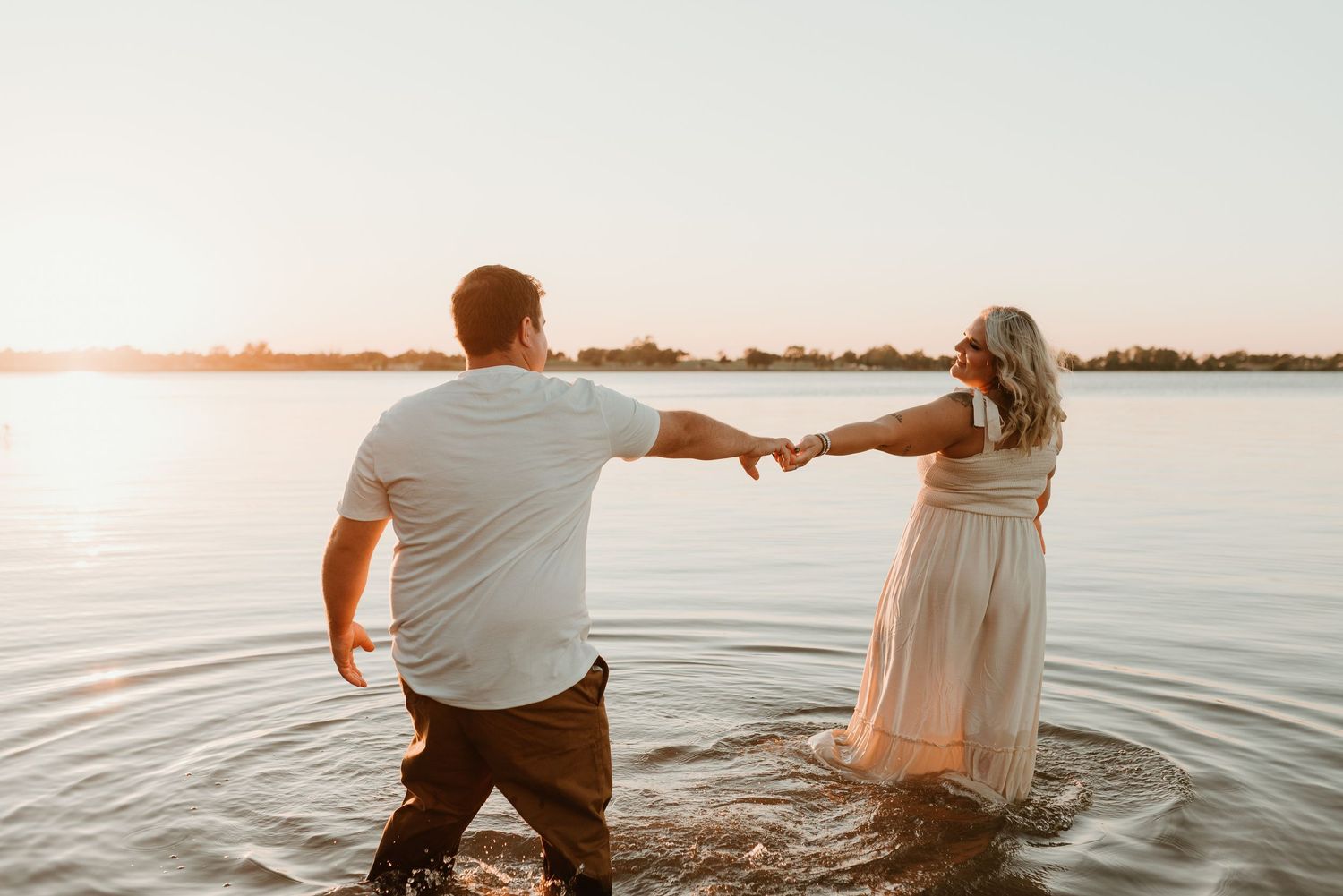 Couple holding hands while wading in a lake at golden hour sunset, romantic outdoor photo.