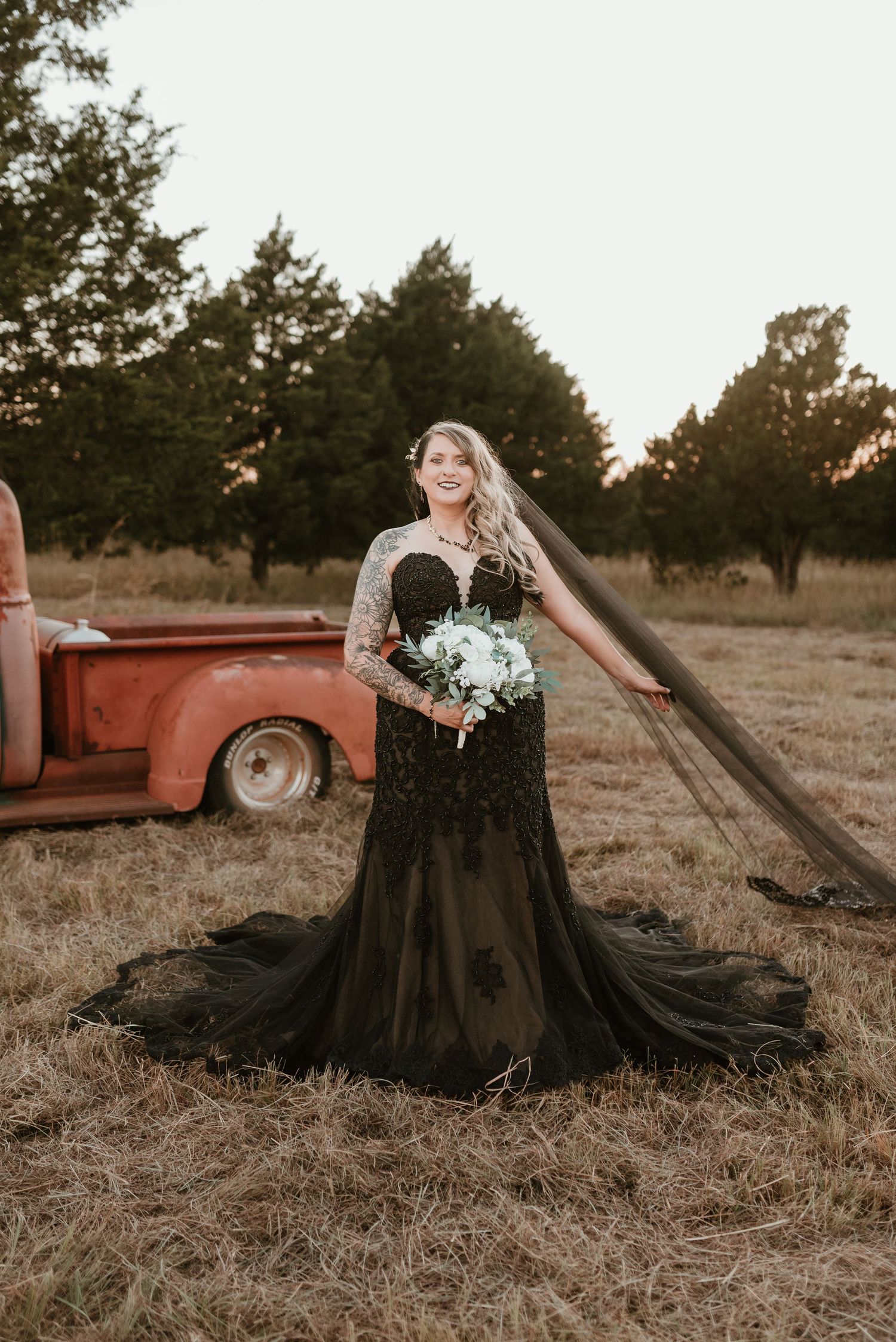 Bride in a stunning black lace wedding gown holding white bouquet in a rustic field at sunset.